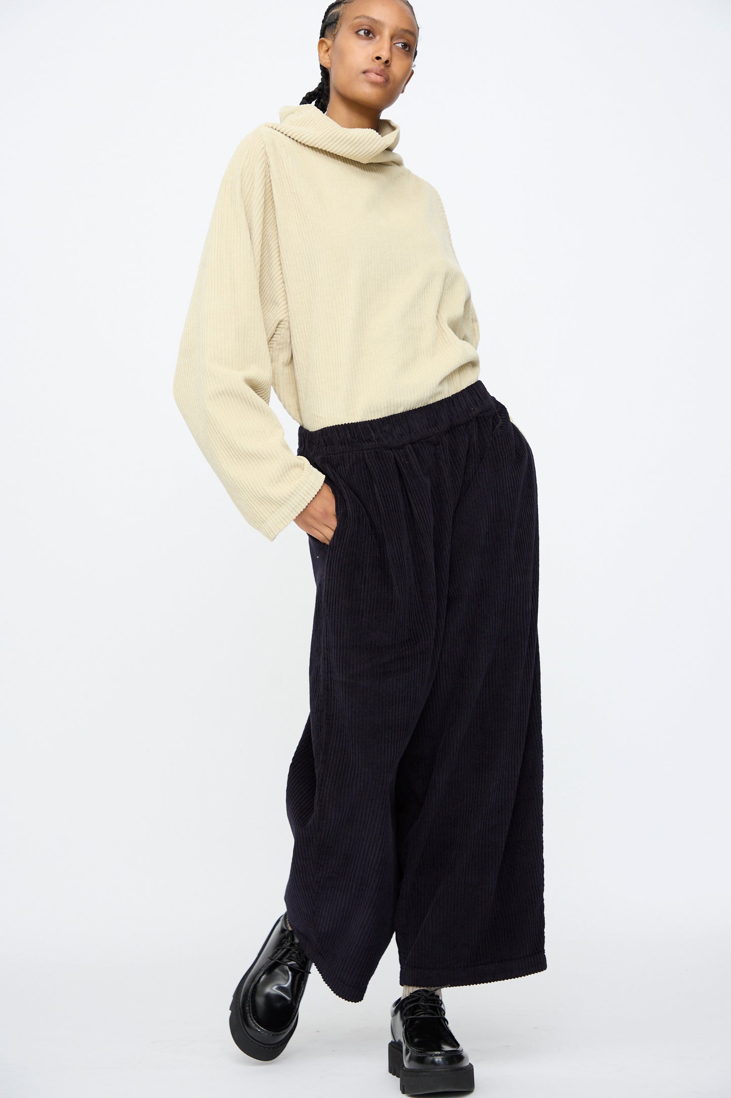 A person stands against a plain white background wearing a loose beige turtleneck sweater, Black Crane Wide Pants in Dark Navy with an elastic waist, and black platform shoes.