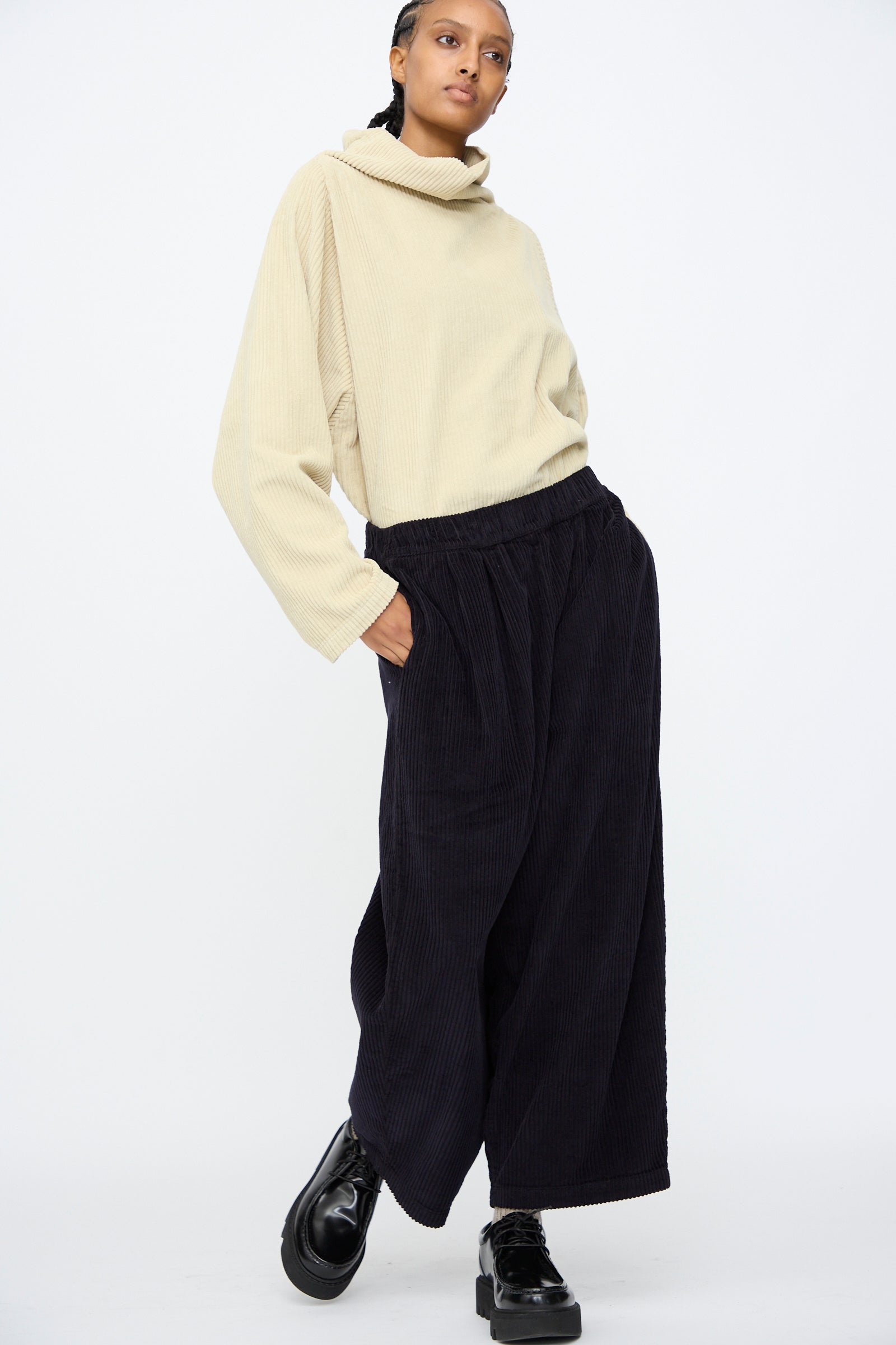 A person stands against a plain white background wearing a loose beige turtleneck sweater, Black Crane Wide Pants in Dark Navy with an elastic waist, and black platform shoes.