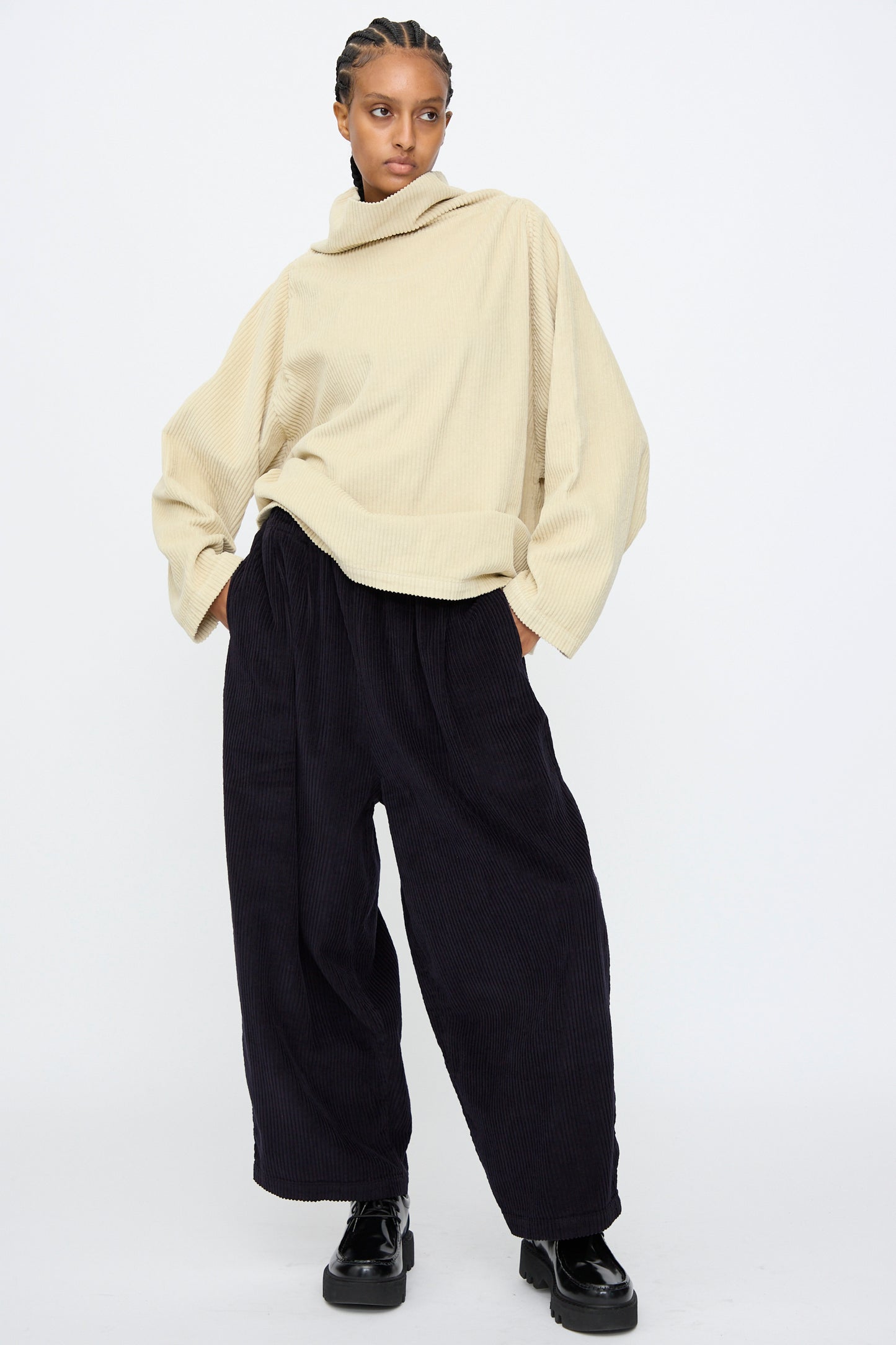 A person stands against a plain background, wearing a loose beige turtleneck sweater, Black Crane Wide Pants in Dark Navy with an elastic waist, and black platform shoes.