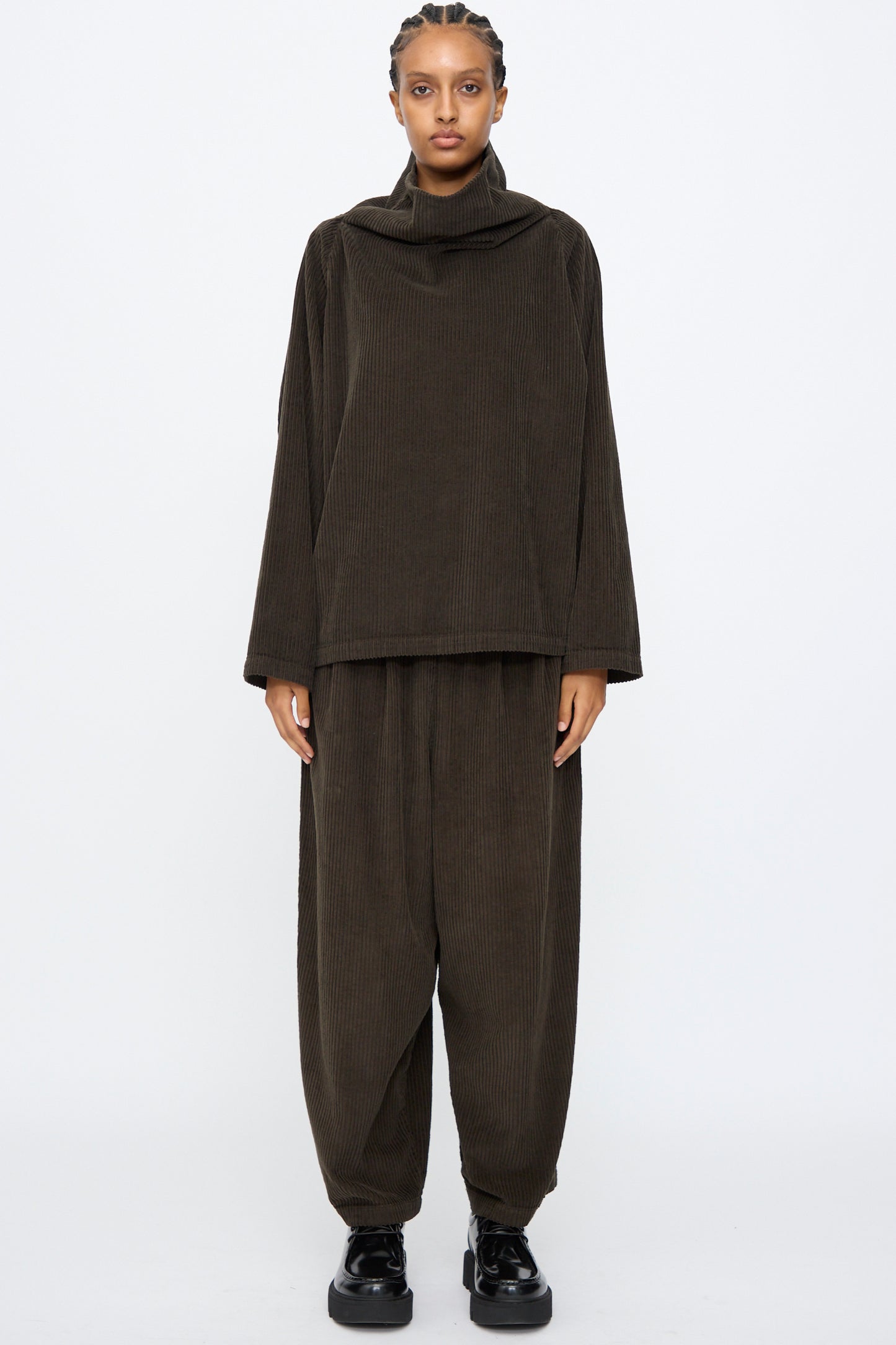 A person faces forward in Black Crane's Wide Pants in Forest, featuring wide-leg corduroy and an elastic waist, styled with a loose dark brown long-sleeve top and black shoes against a plain light background.
