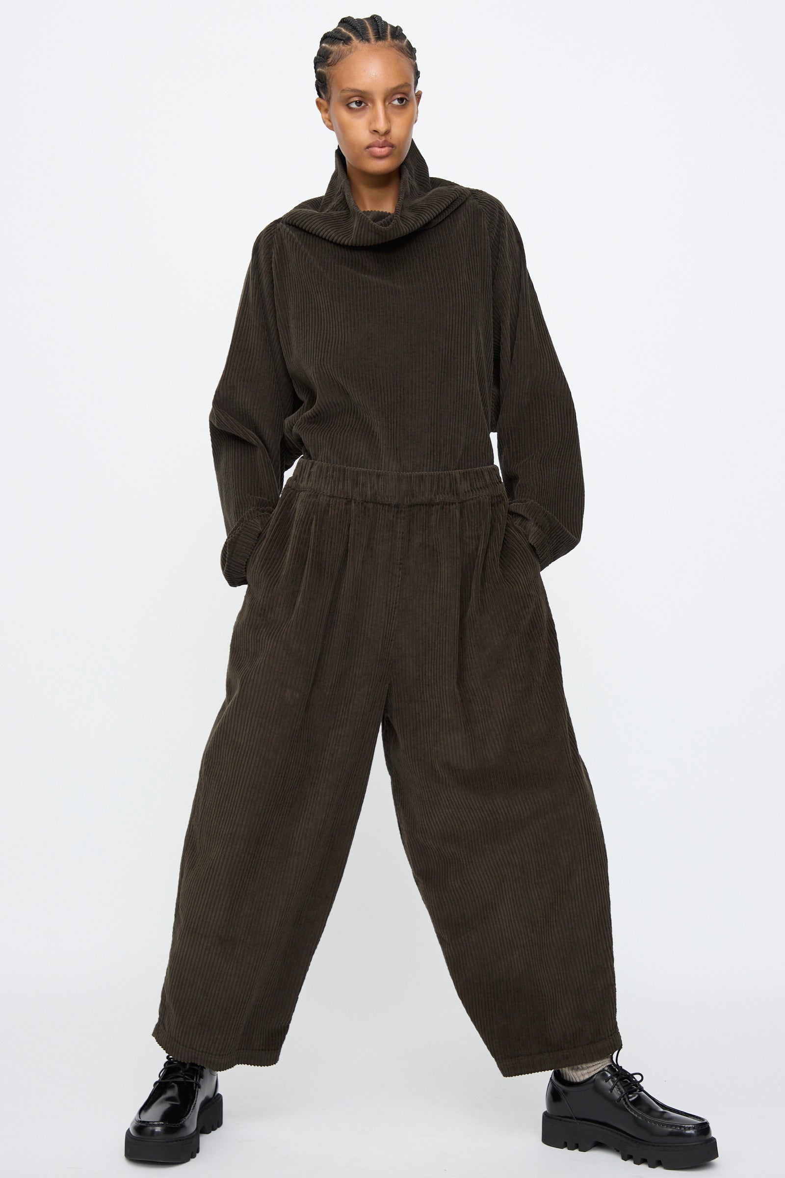 A person stands with hands in pockets, wearing Black Crane's Wide Pants in Forest—loose-fitting corduroy pants with an elastic waist—paired with a dark high-collar top and black chunky shoes, set against a plain background.
