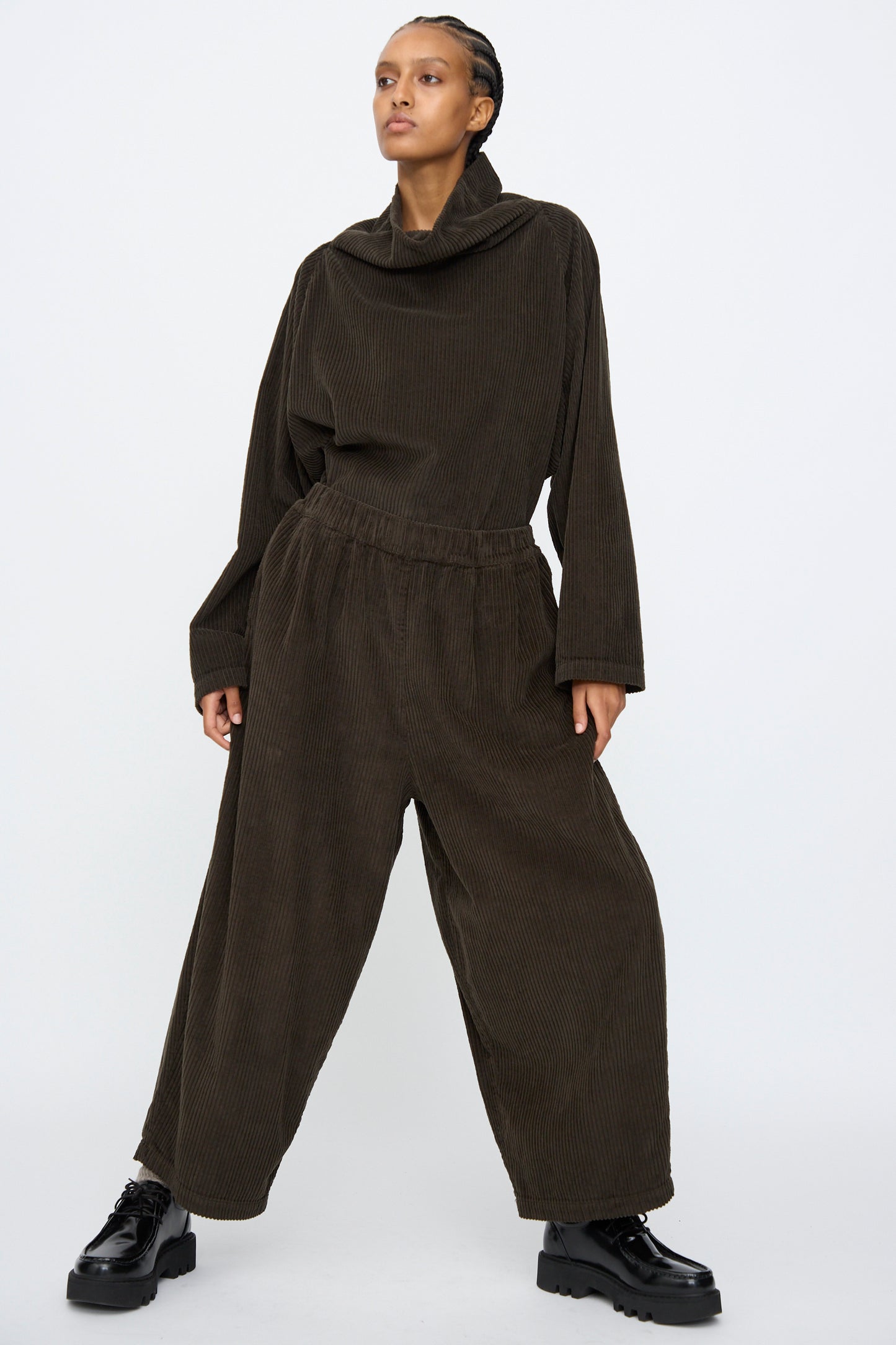 A person stands against a plain background wearing Black Crane’s Wide Pants in Forest with an elastic waist, paired with a loose cowl-neck top and black platform shoes. This stylish outfit is made in the USA.