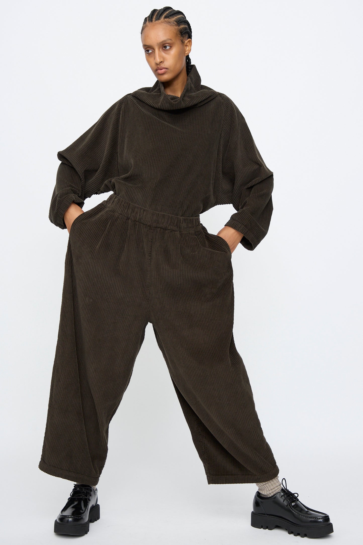 A person stands with hands in pockets, wearing Black Crane's Wide Pants in Forest—a loose-fitting, dark brown corduroy style—paired with black shoes against a plain white background.