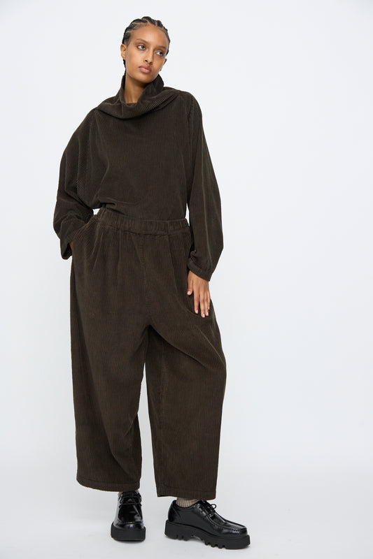 A person stands against a plain background wearing Black Crane's Wide Pants in Forest, a loose-fitting corduroy style with an elastic waist, paired with black platform shoes.