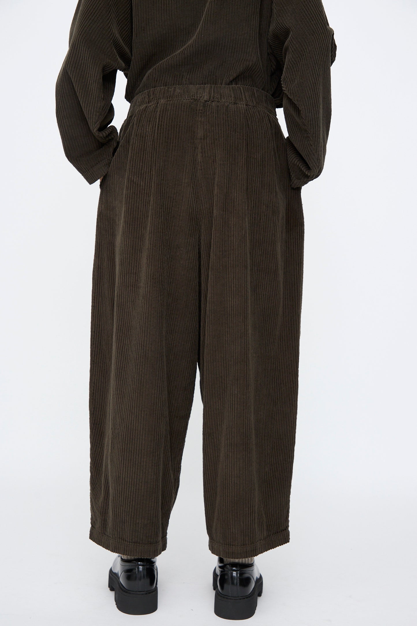 Person wearing Black Crane's Wide Pants in Forest—loose-fitting, wide-leg corduroy with an elastic waist—paired with black shoes, photographed from behind against a plain white background.