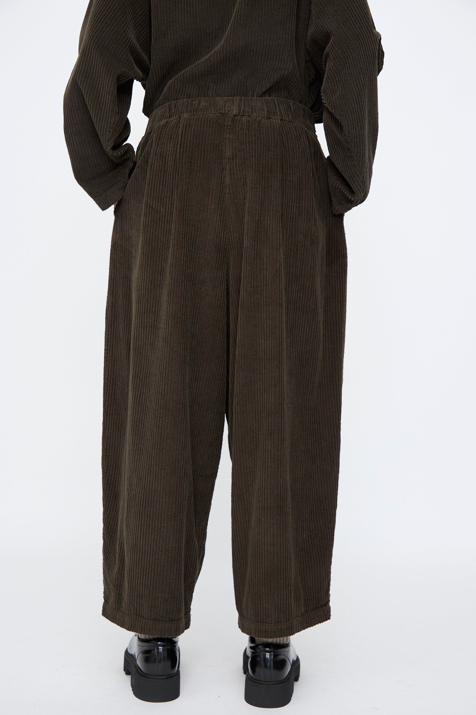 Person wearing Black Crane's Wide Pants in Forest—loose-fitting, wide-leg corduroy with an elastic waist—paired with black shoes, photographed from behind against a plain white background.