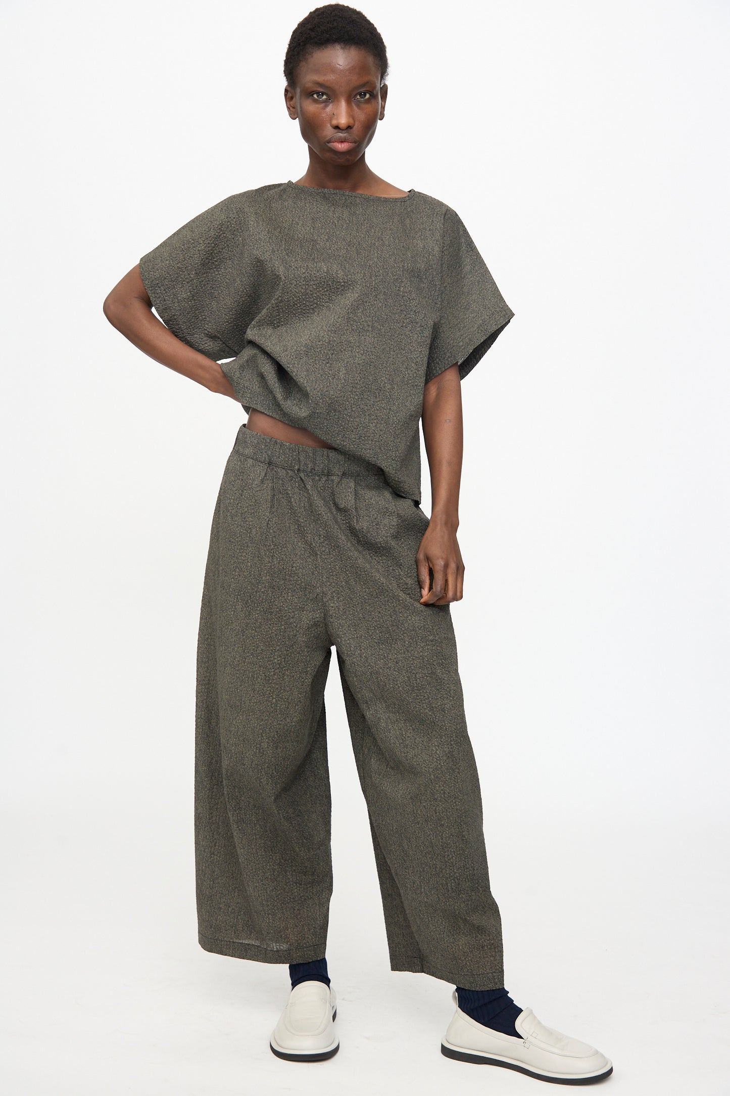 Person standing against a plain background wearing a relaxed fit grey top, Black Crane Wide Pants with Micro Wave Texture in Steel, navy socks, and white slip-on shoes.