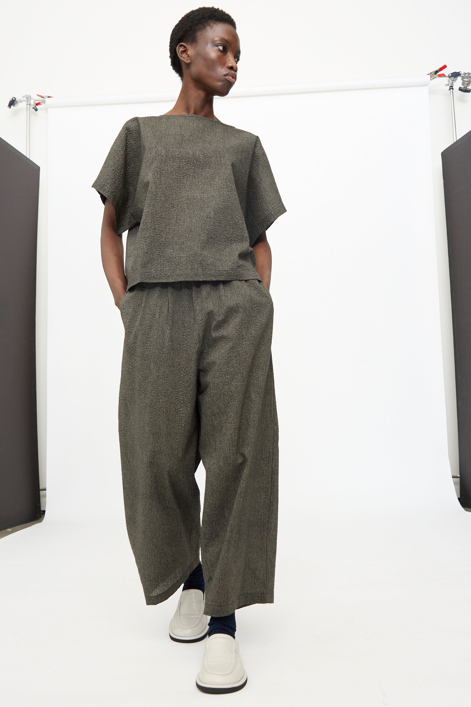 A person wears Black Crane's Wide Pants with Micro Wave Texture in Steel, paired with a boxy gray short-sleeve top and white shoes, standing against a plain white background.