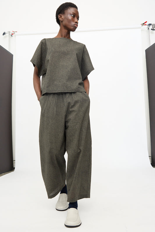 A person wears Black Crane's Wide Pants with Micro Wave Texture in Steel, paired with a boxy gray short-sleeve top and white shoes, standing against a plain white background.