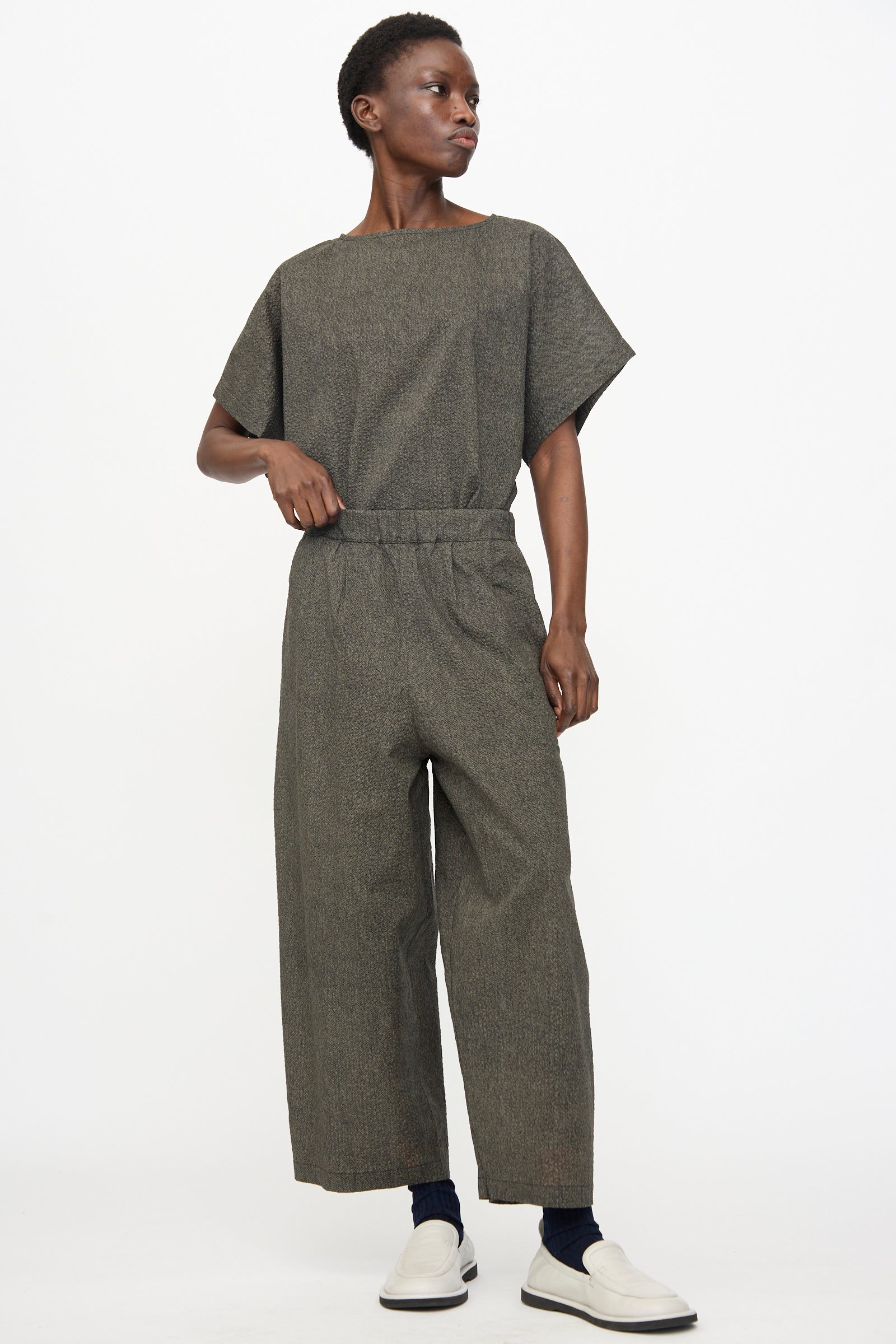 A person stands against a white background wearing Black Crane’s Wide Pants with Micro Wave Texture in Steel, a grey relaxed-fit short-sleeve top, white slip-on shoes, and dark socks.