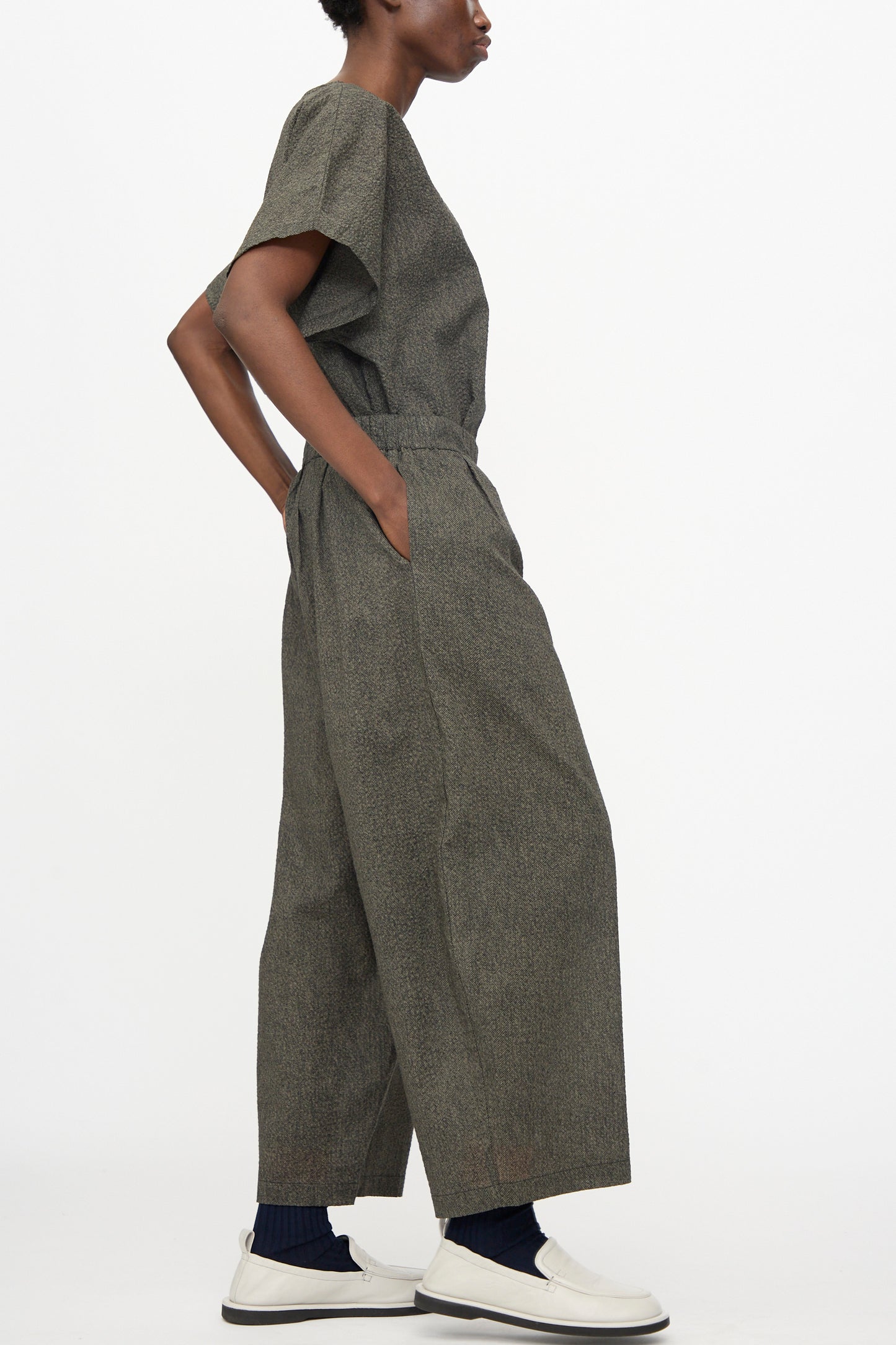 A person in profile wears Black Crane's Wide Pants with Micro Wave Texture in Steel, styled with a relaxed gray Japanese cotton short-sleeve top, white slip-on shoes, and dark socks.