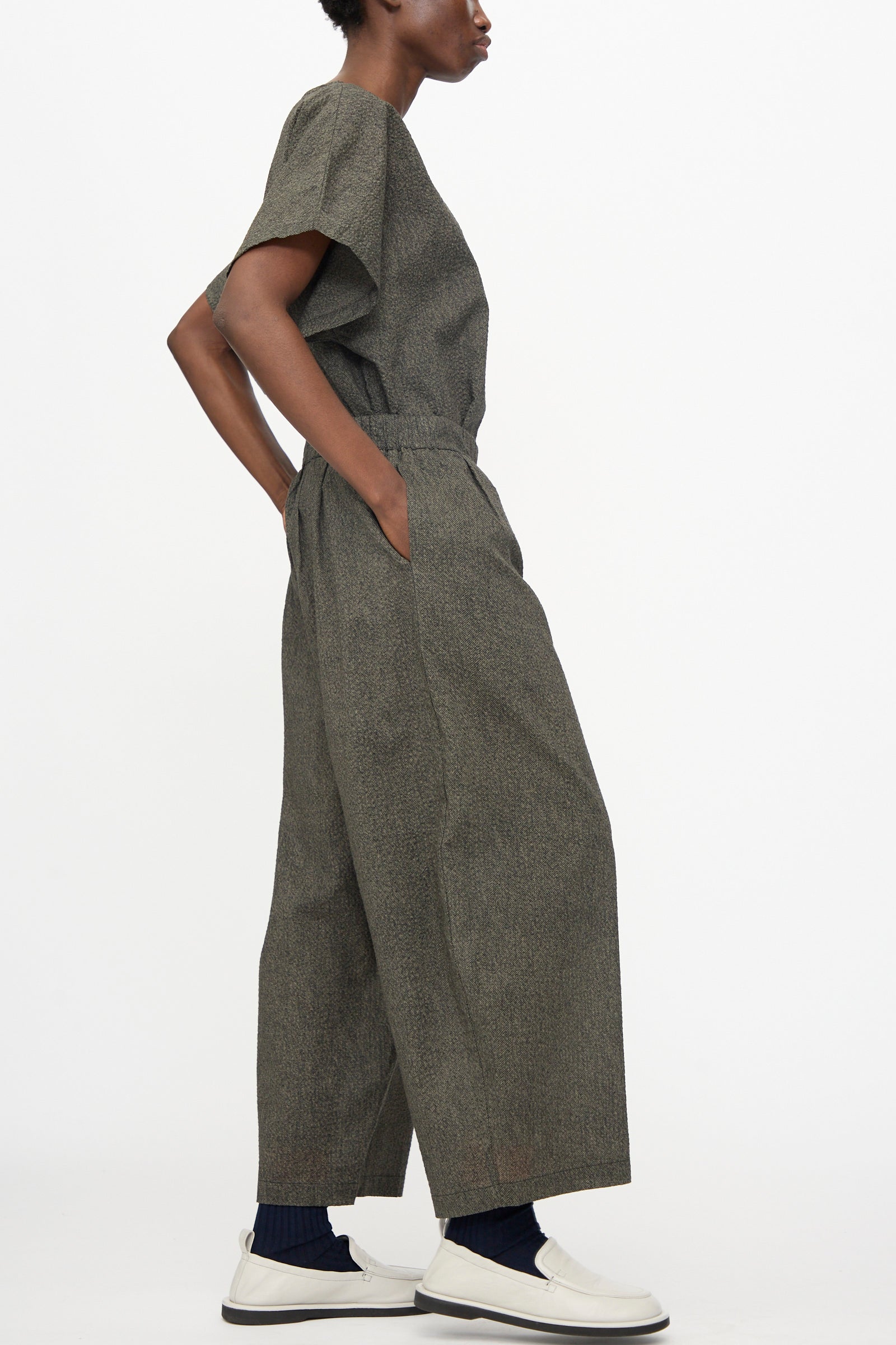 A person in profile wears Black Crane's Wide Pants with Micro Wave Texture in Steel, styled with a relaxed gray Japanese cotton short-sleeve top, white slip-on shoes, and dark socks.