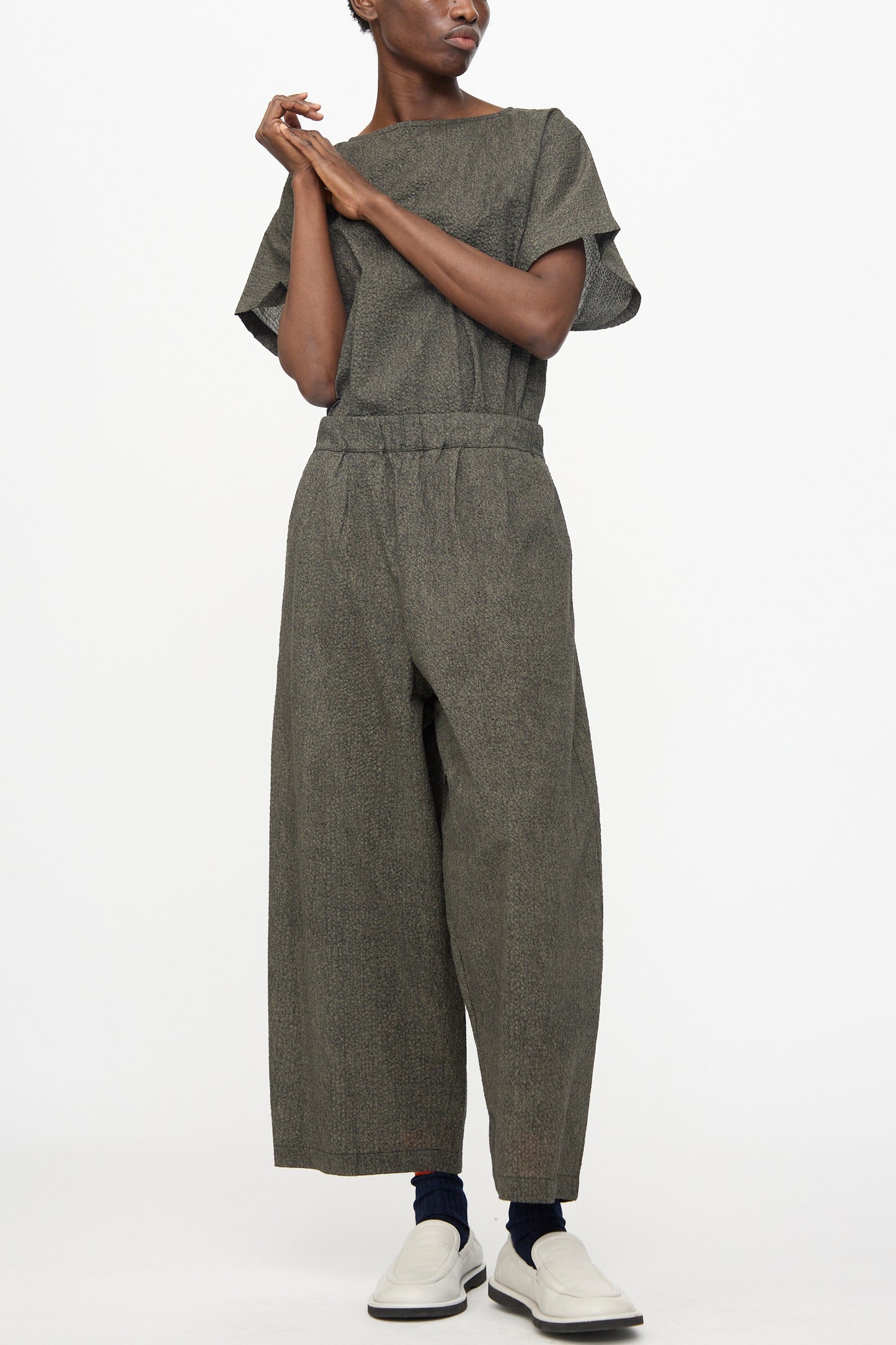 A person wearing Black Crane’s Wide Pants with Micro Wave Texture in Steel, paired with black socks and white slip-on shoes, stands against a plain white background.