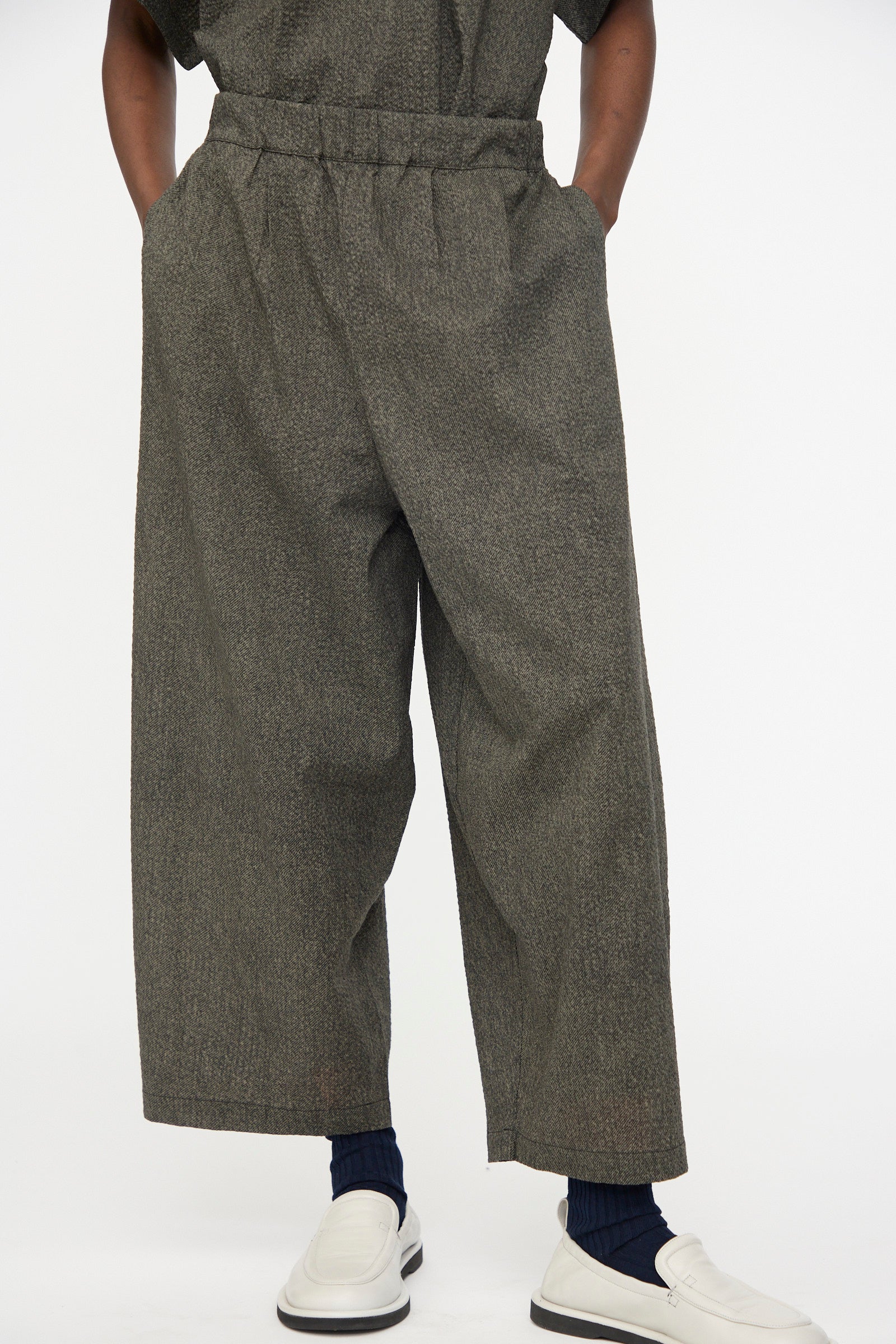 Person wearing Black Crane Wide Pants with Micro Wave Texture in Steel, hands in pockets, navy socks, and white slip-on shoes against a plain background.