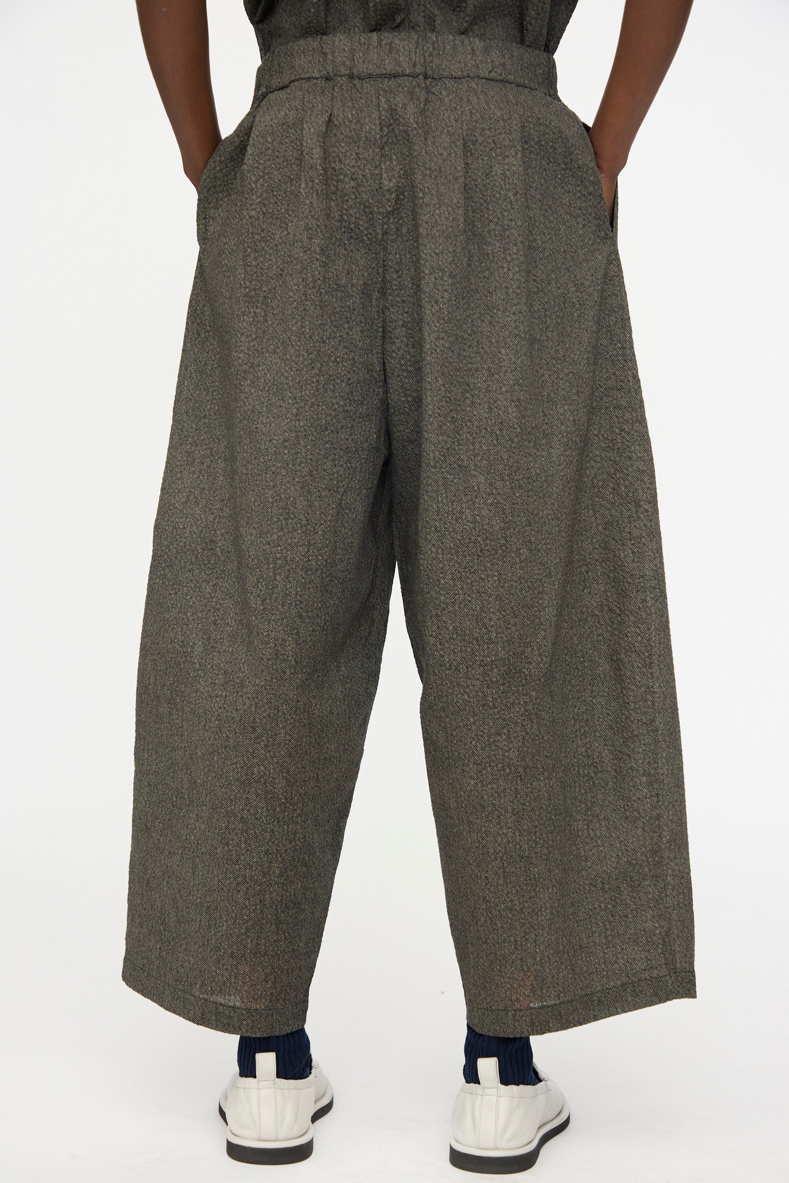 Person wearing Black Crane Wide Pants with Micro Wave Texture in Steel, paired with white sneakers and blue socks, stands hands-in-pockets, photographed from the back.