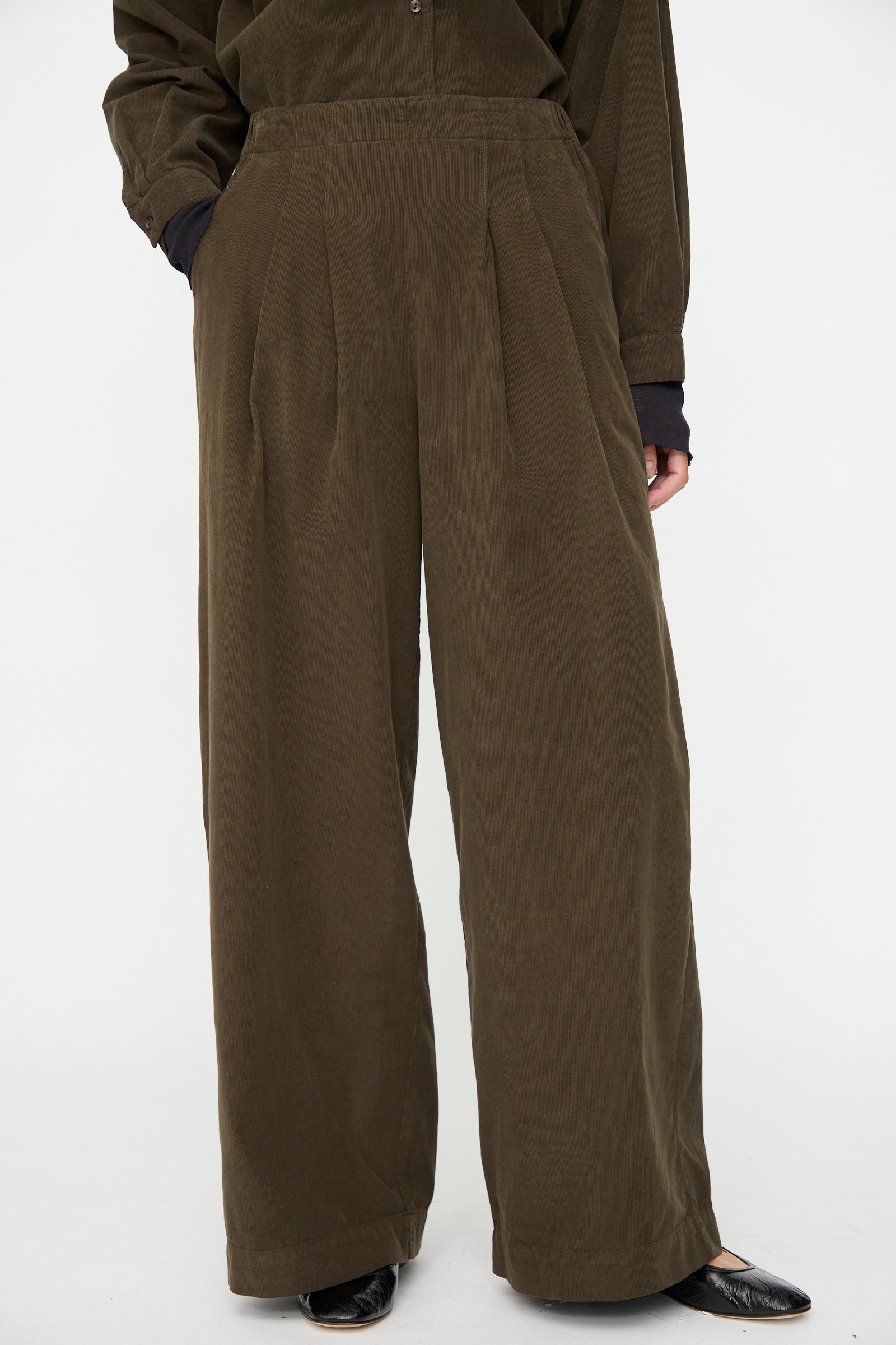 A person wears Black Crane's Wide Pleated Pants in Dark Forest—relaxed fit, high-waisted, wide-leg—with long sleeves and black pointed shoes, standing against a plain white background.