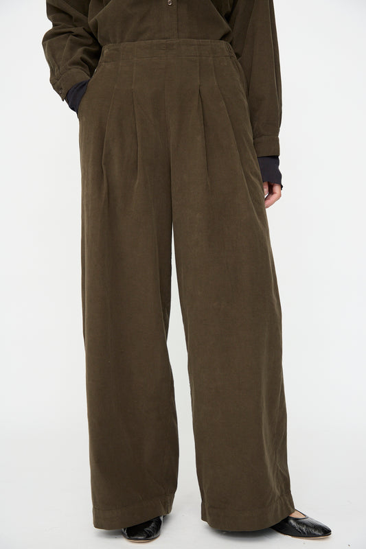 A person wears Black Crane's Wide Pleated Pants in Dark Forest—relaxed fit, high-waisted, wide-leg—with long sleeves and black pointed shoes, standing against a plain white background.