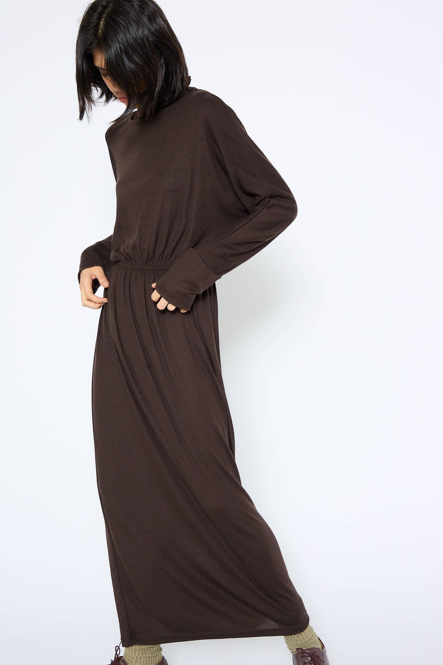 A person with shoulder-length dark hair wears the Wool I-Dress in Cacao by Black Crane, a relaxed fit, long-sleeved, ankle-length brown maxi dress made of Japanese wool jersey, paired with gray socks and brown shoes, against a plain white background.