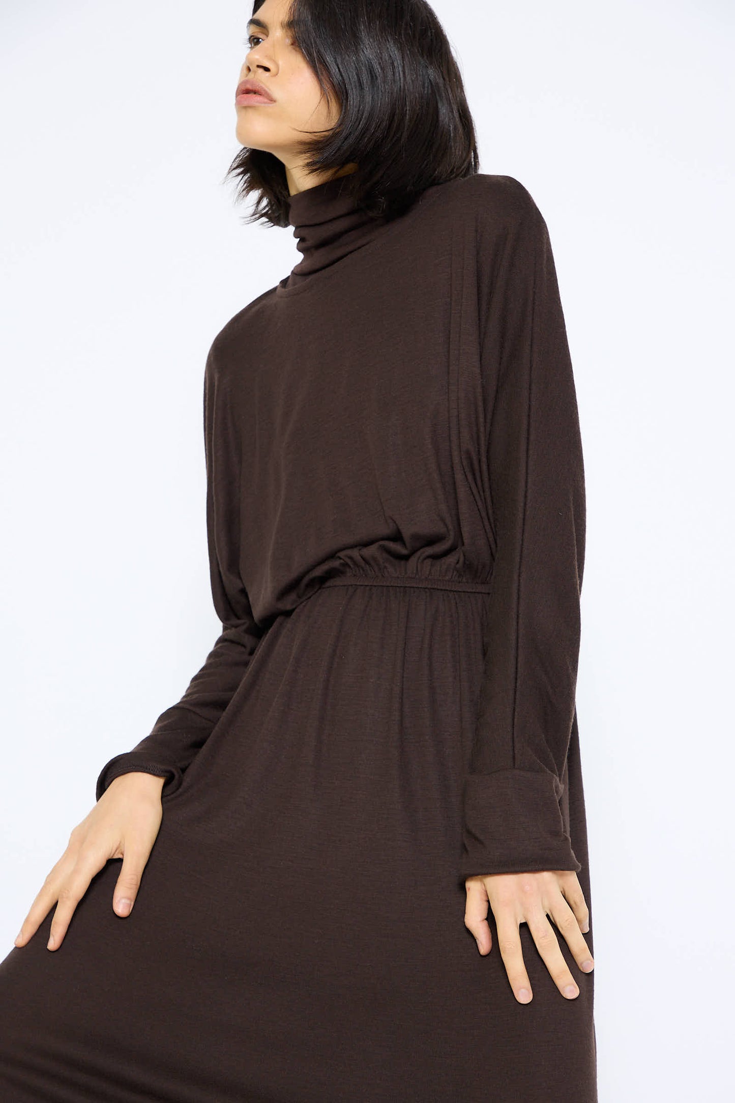 Person with shoulder-length dark hair wears the Wool I-Dress in Cacao from Black Crane, a long-sleeved dark brown turtleneck maxi dress made of Japanese wool jersey, posing against a light background.