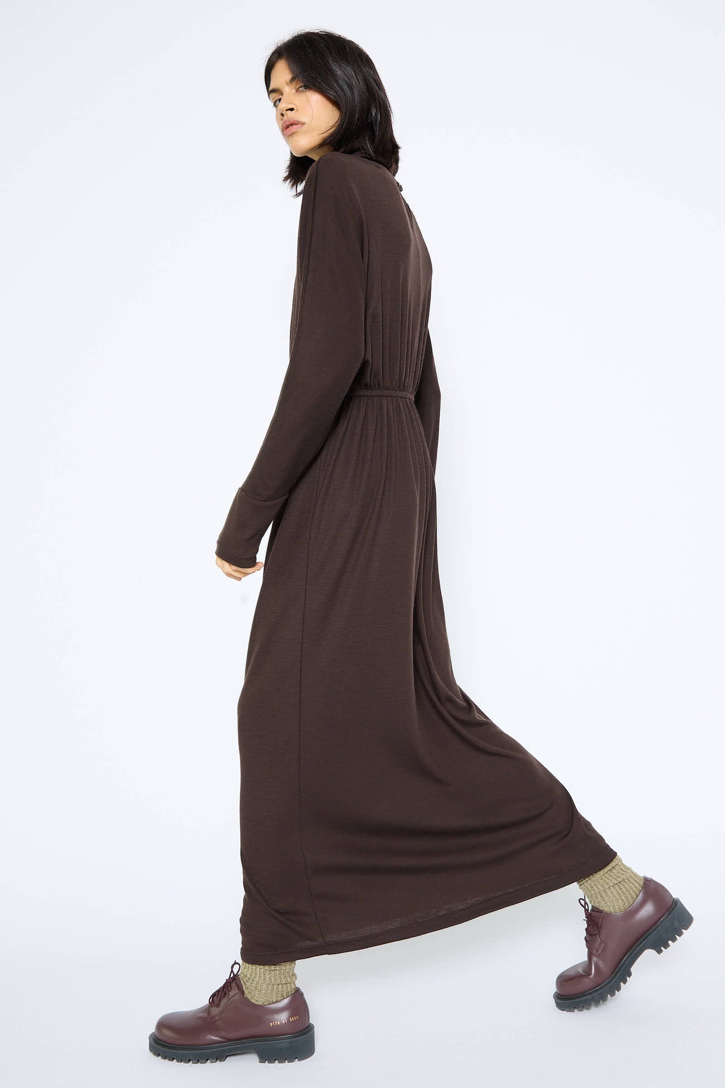 A person wearing a relaxed fit, long Wool I-Dress in Cacao by Black Crane made of Japanese wool jersey and burgundy boots with green socks is walking against a plain white background.