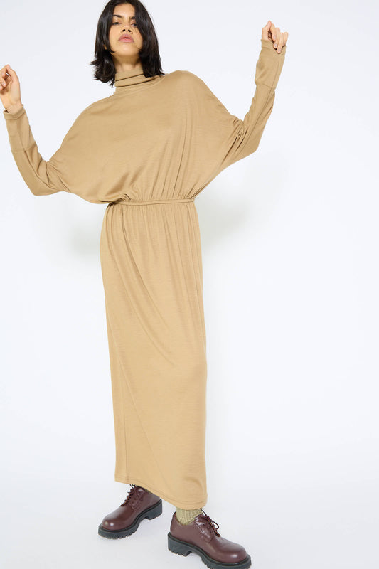 A person poses in the Wool I-Dress in Camel by Black Crane, a tan long-sleeve, ankle-length maxi dress with a cinched waist and brown lace-up boots. Made from Japanese wool jersey, the dress drapes elegantly as they stand against a plain white background.