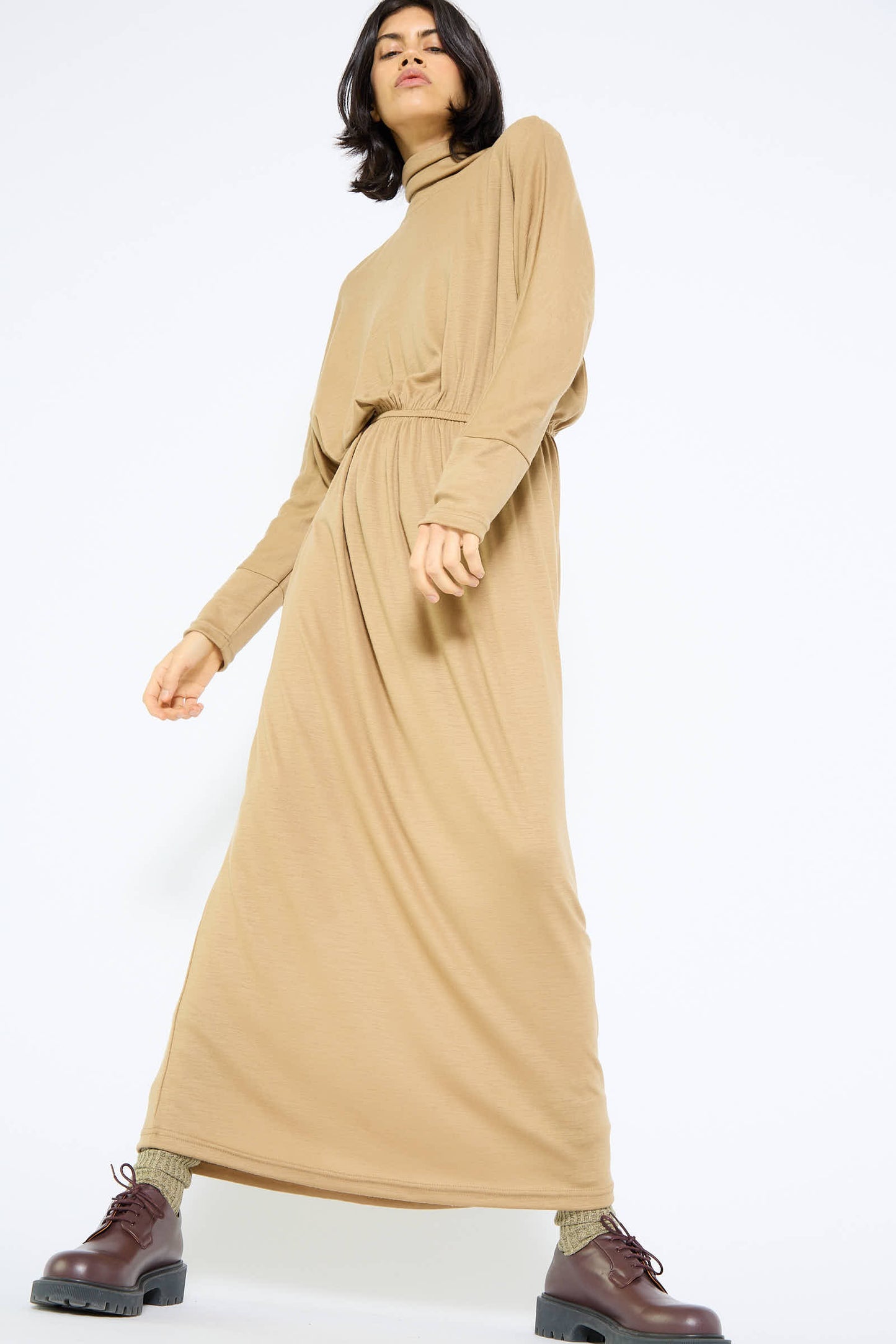 A person wearing the Wool I-Dress in Camel by Black Crane, featuring long dolman sleeves, pairs it with brown boots while posing against a plain white background.