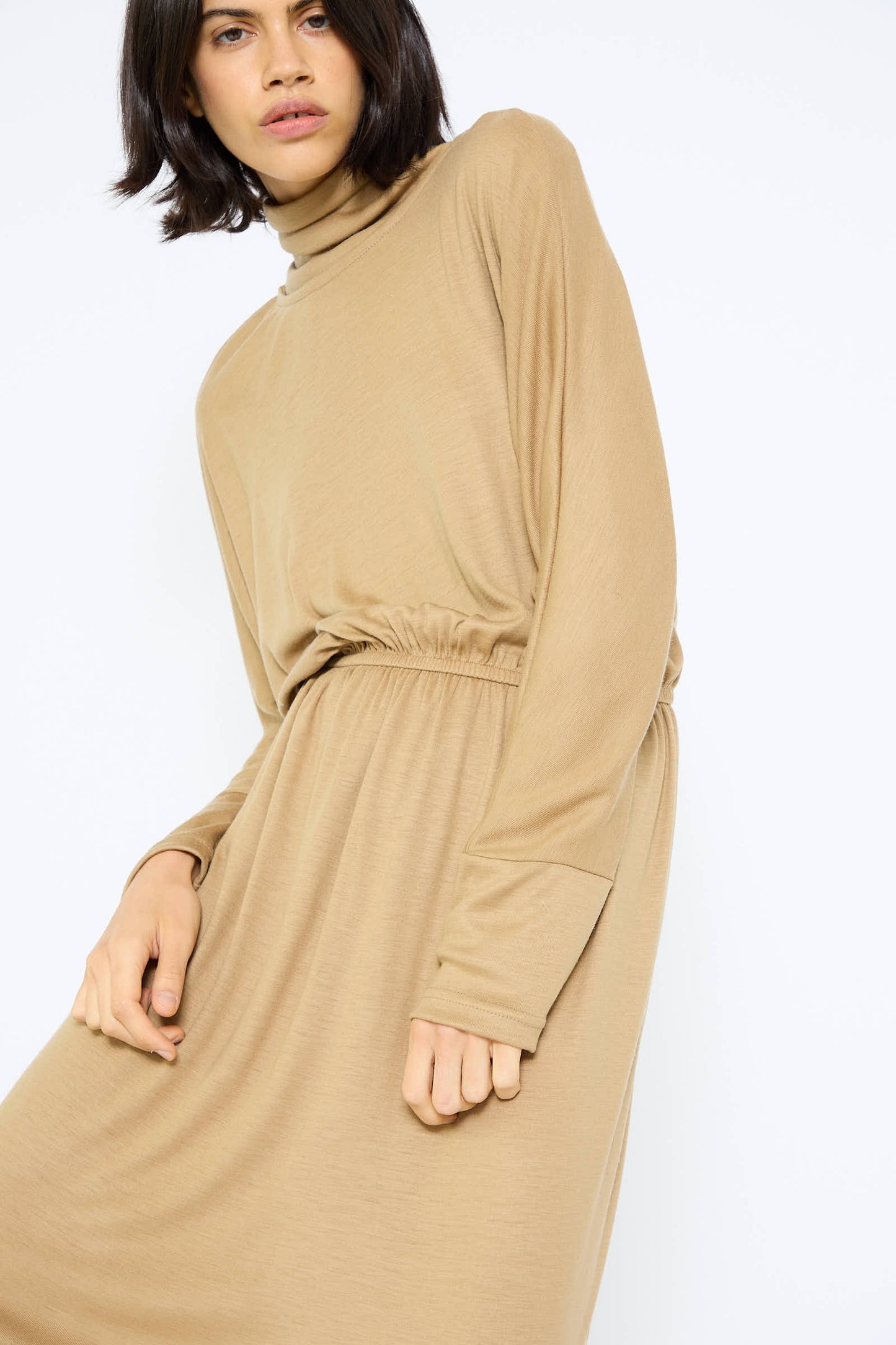 A person with shoulder-length dark hair wears the Wool I-Dress in Camel by Black Crane, featuring a loose fit, long dolman sleeves, and a cinched waist.