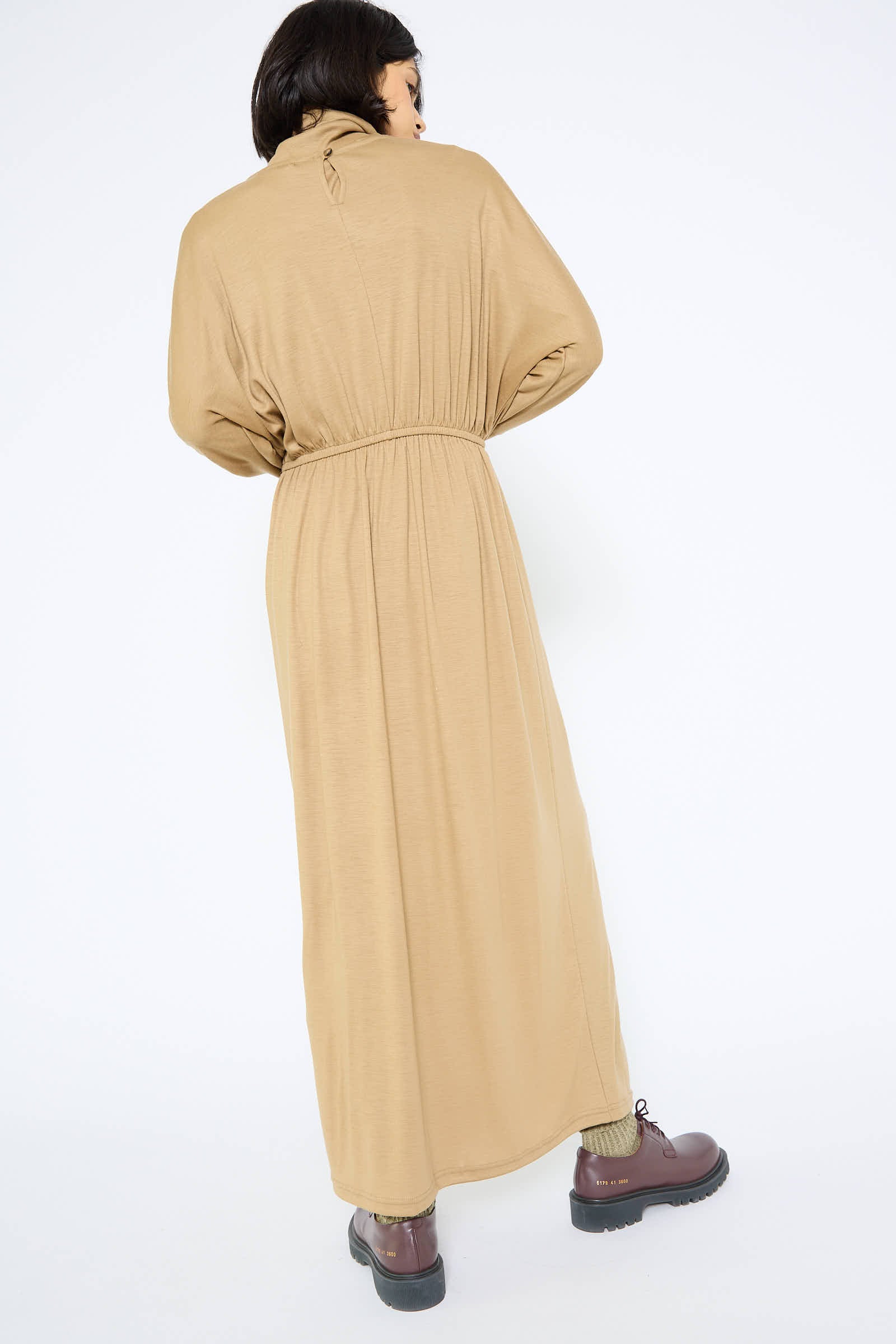 Person standing with their back to the camera, wearing the Black Crane Wool I-Dress in Camel with gathered waist details and brown shoes. The dress features long sleeves and a high neckline.