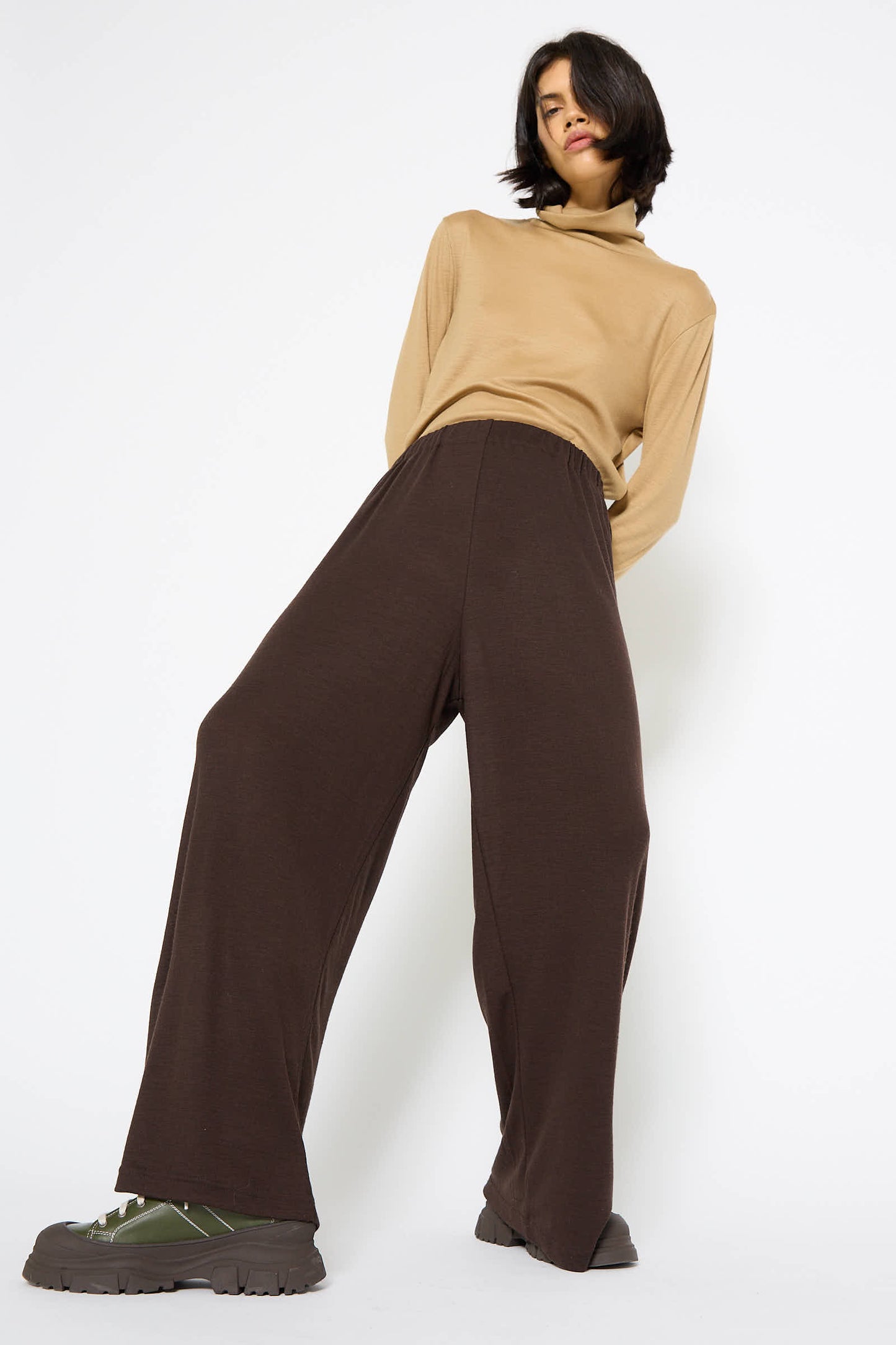 Person standing against a plain white background, wearing a beige Japanese wool jersey turtleneck sweater and Wool Straight Pant in Cacao by Black Crane with an elasticated waist. They have chunky green and brown shoes and shoulder-length dark hair.