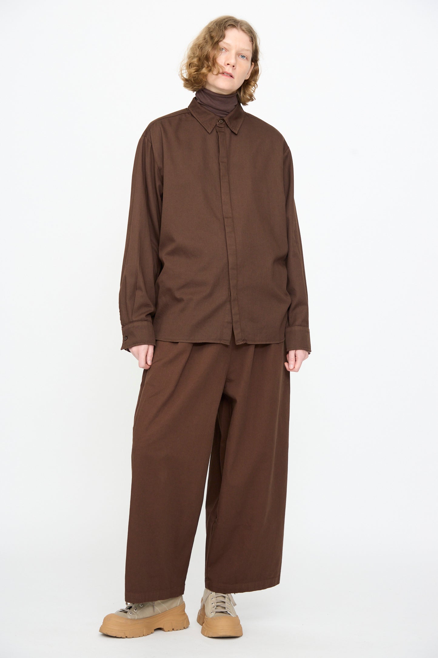 A person wears Black Crane’s Work Shirt in Dark Cacao, styled with matching wide-leg pants, tan shoes, and a brown turtleneck, standing against a plain white background.