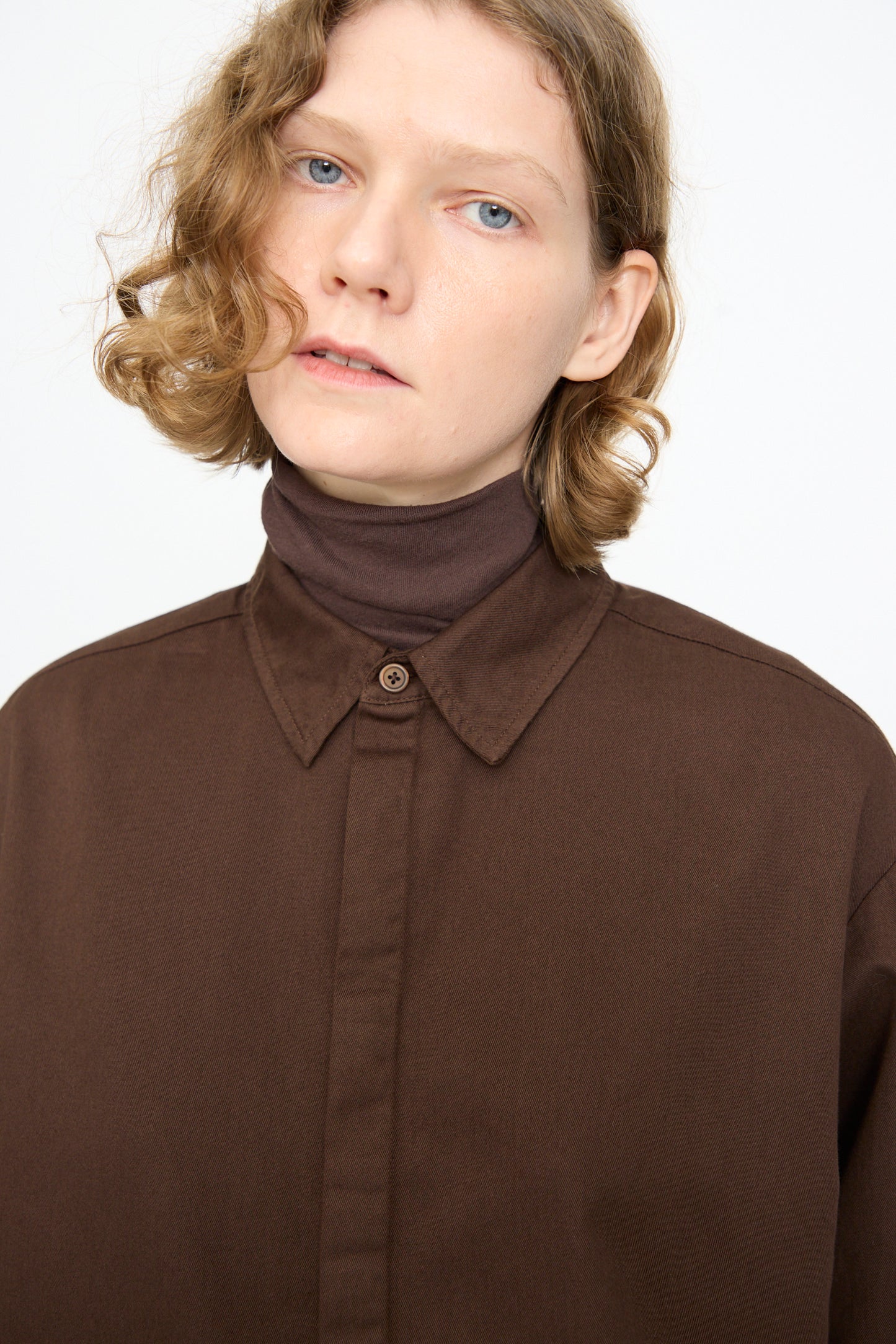 A person with short, wavy blond hair wears the Black Crane Work Shirt in Dark Cacao—made in the USA from cotton—layered over a brown turtleneck, looking slightly upward against a plain white background.
