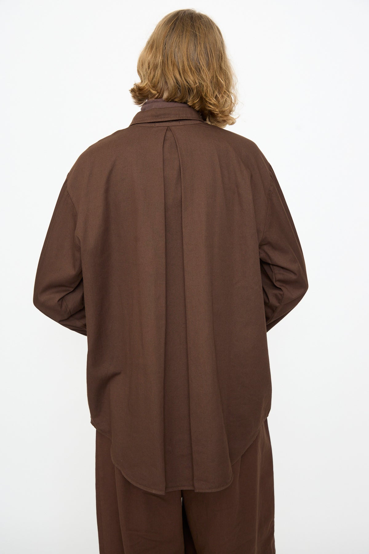 A person with shoulder-length hair, shown from behind, is wearing the Black Crane Work Shirt in Dark Cacao along with matching brown pants against a plain white background.