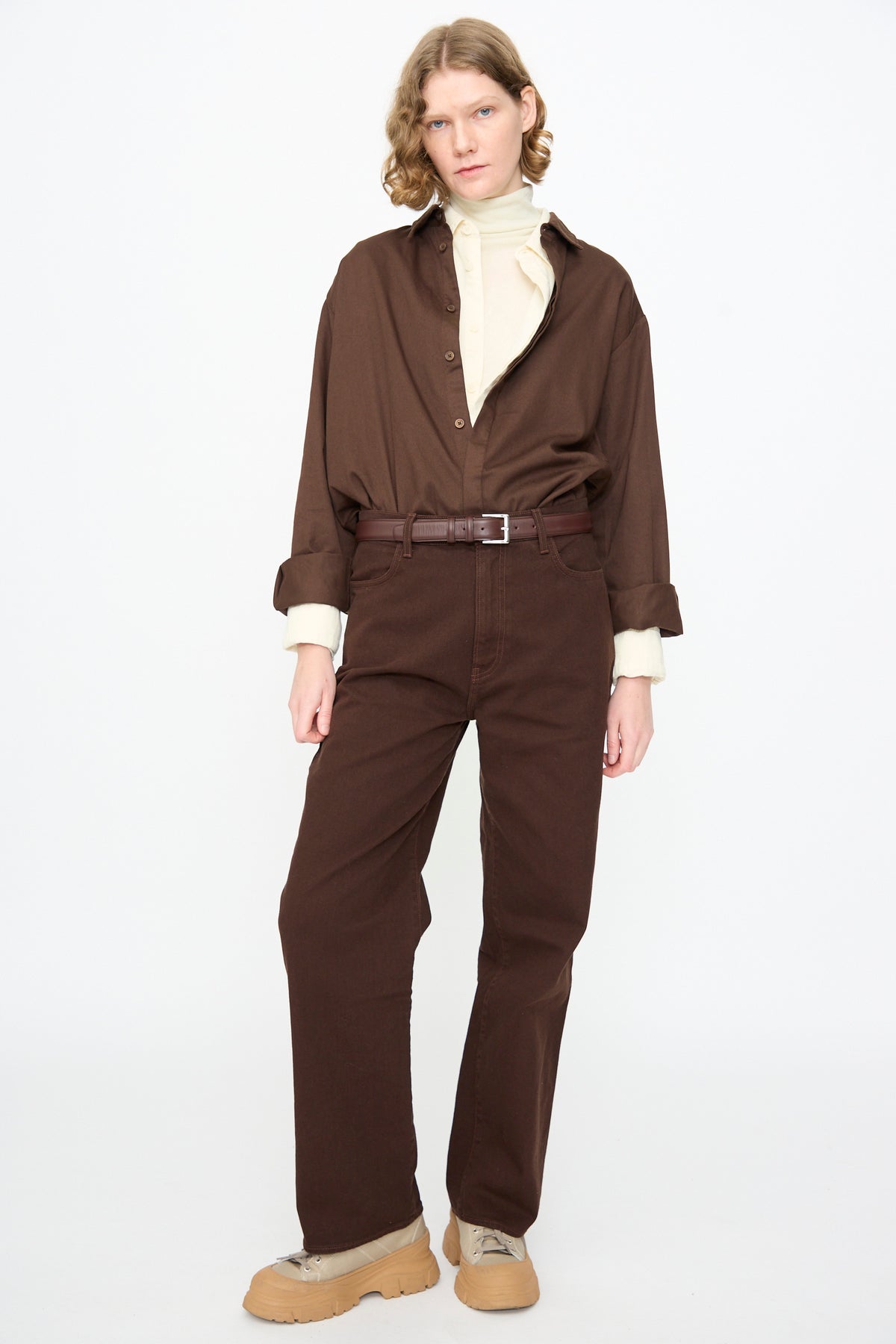 A person stands against a plain background, wearing a beige turtleneck, brown pants, tan shoes, and the Work Shirt in Dark Cacao by Black Crane.