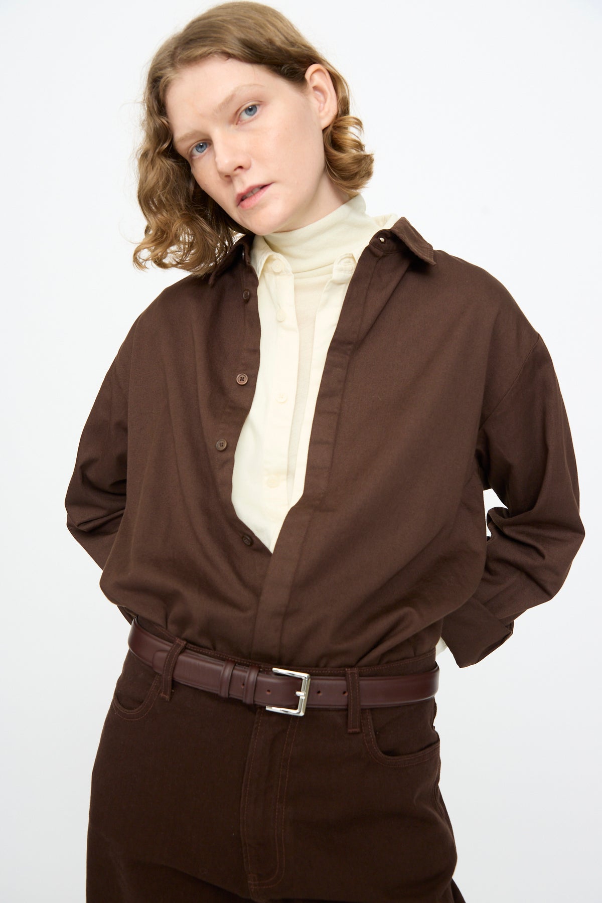 A person with short wavy hair is standing against a plain white background, wearing the Black Crane Work Shirt in Dark Cacao—a relaxed-fit cotton piece made in the USA—layered over a white turtleneck and paired with brown pants and a brown belt for a stylish look.