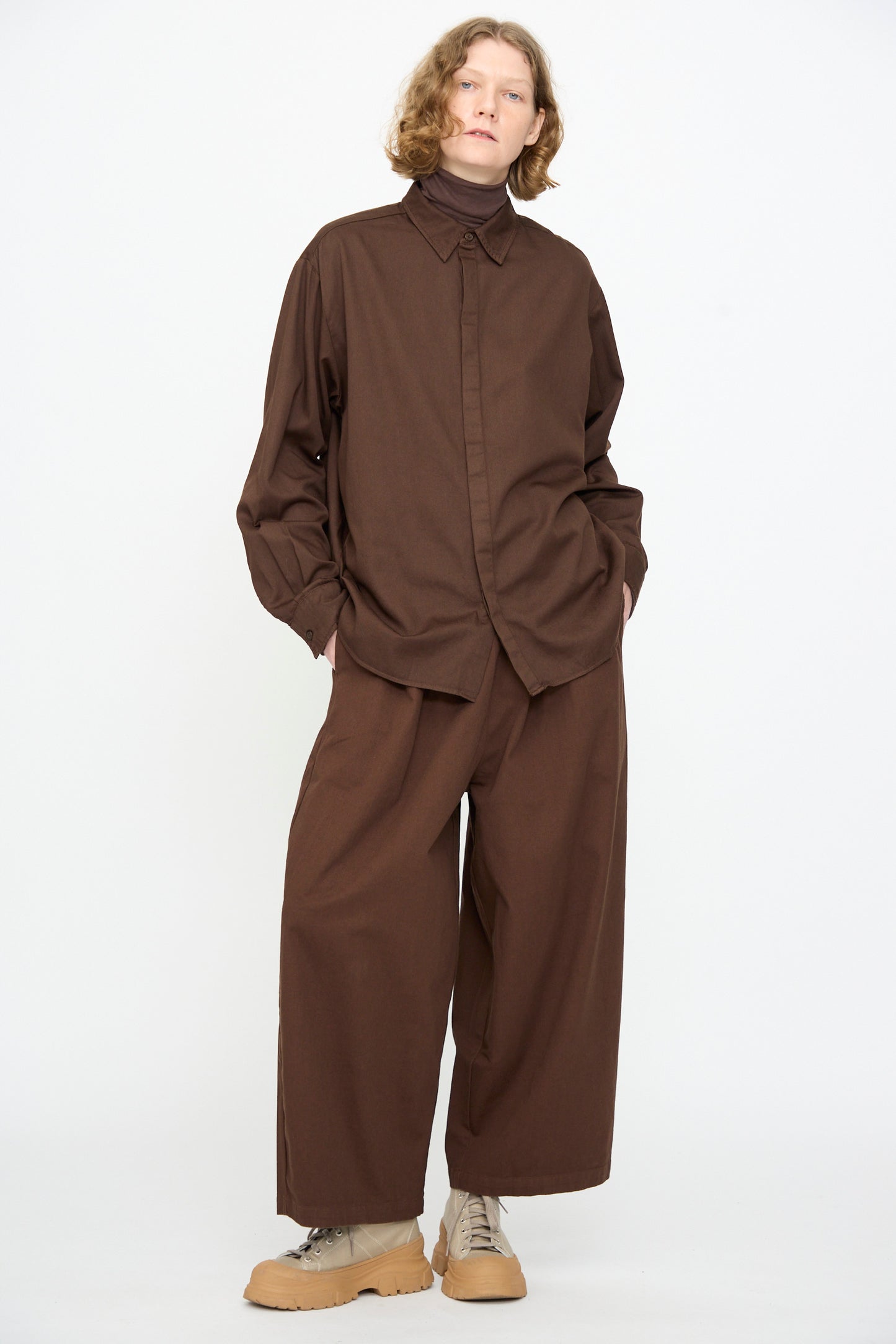 A person wears the Black Crane Work Shirt in Dark Cacao—a loose, dark brown cotton shirt—paired with wide brown pants and light brown shoes, standing against a plain white background. Made in the USA, this relaxed fit shirt combines comfort with timeless style.