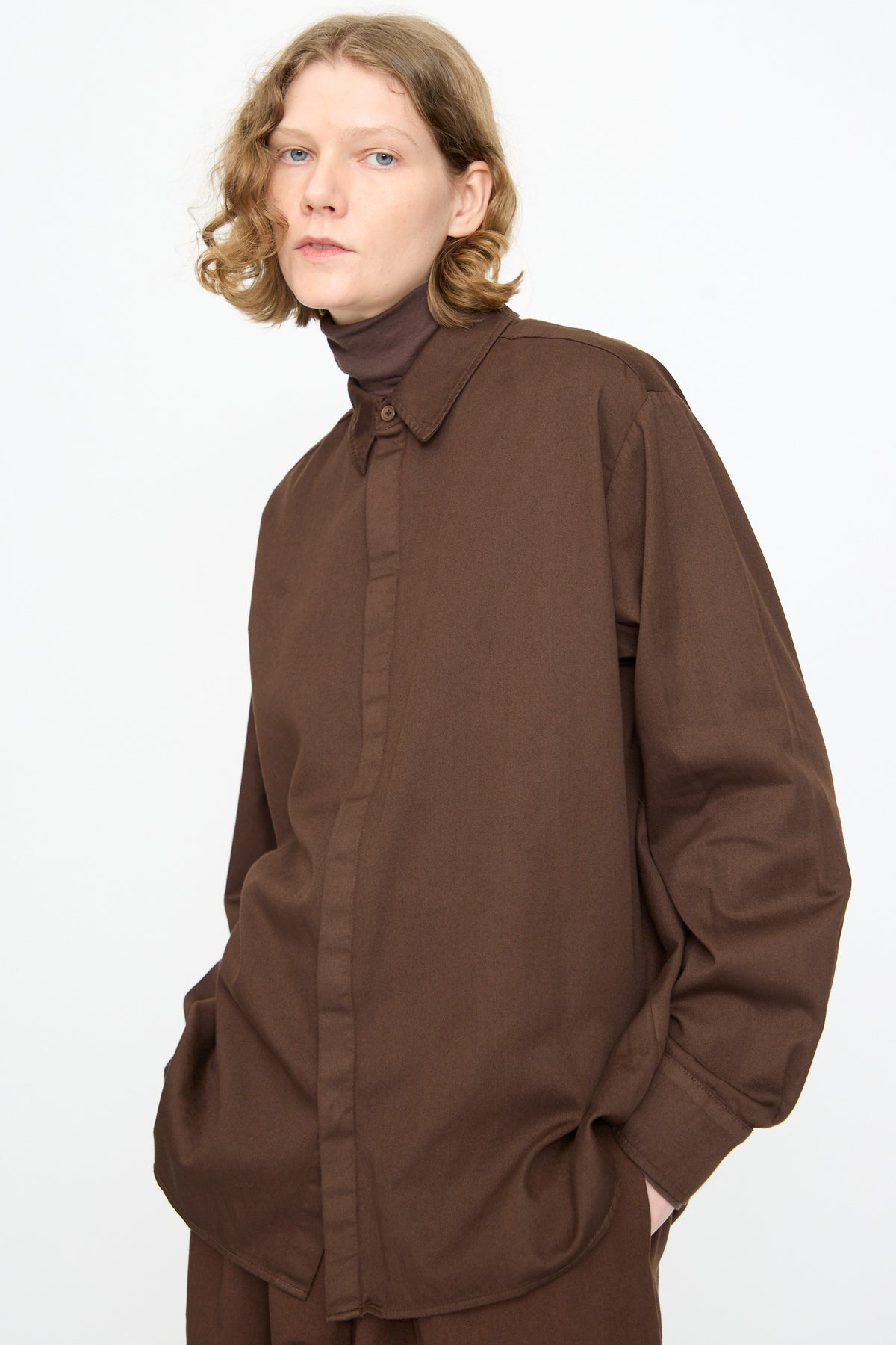 A person with shoulder-length blond hair stands against a plain white background, one hand in their pocket, wearing the Black Crane Work Shirt in Dark Cacao—a relaxed fit, loose brown button-up—layered over a dark brown turtleneck.