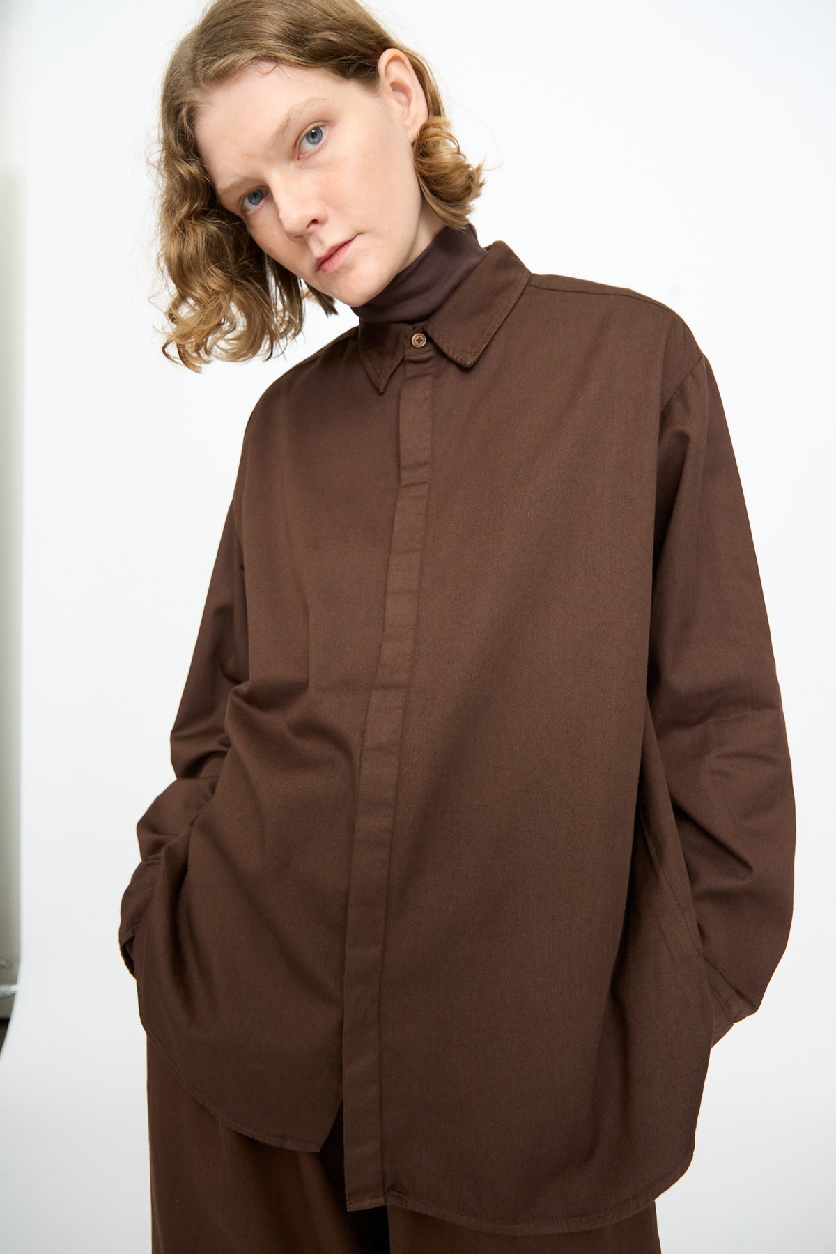 A person with short, wavy blonde hair is wearing the Black Crane Work Shirt in Dark Cacao—a relaxed fit, dark brown cotton shirt made in the USA—with their hands in their pockets, standing against a plain white background.