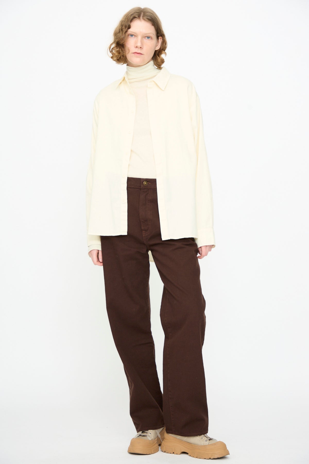 A person wearing Black Crane’s Work Shirt in Natural, layered over a cream turtleneck, paired with brown wide-leg pants and tan sneakers, standing against a plain white background.