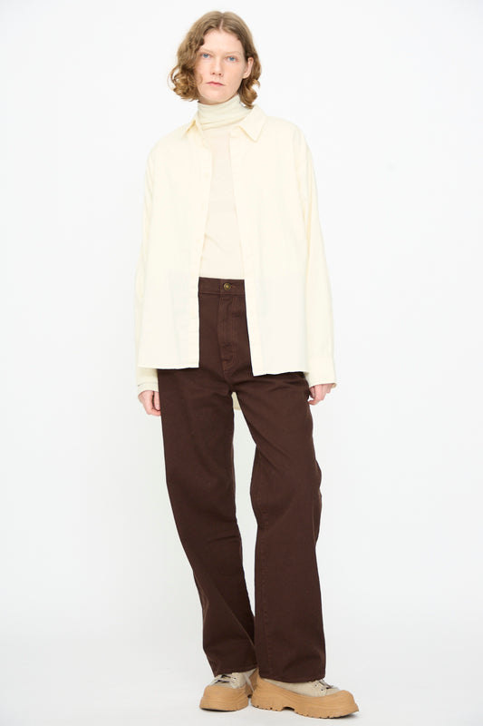 A person wearing Black Crane’s Work Shirt in Natural, layered over a cream turtleneck, paired with brown wide-leg pants and tan sneakers, standing against a plain white background.