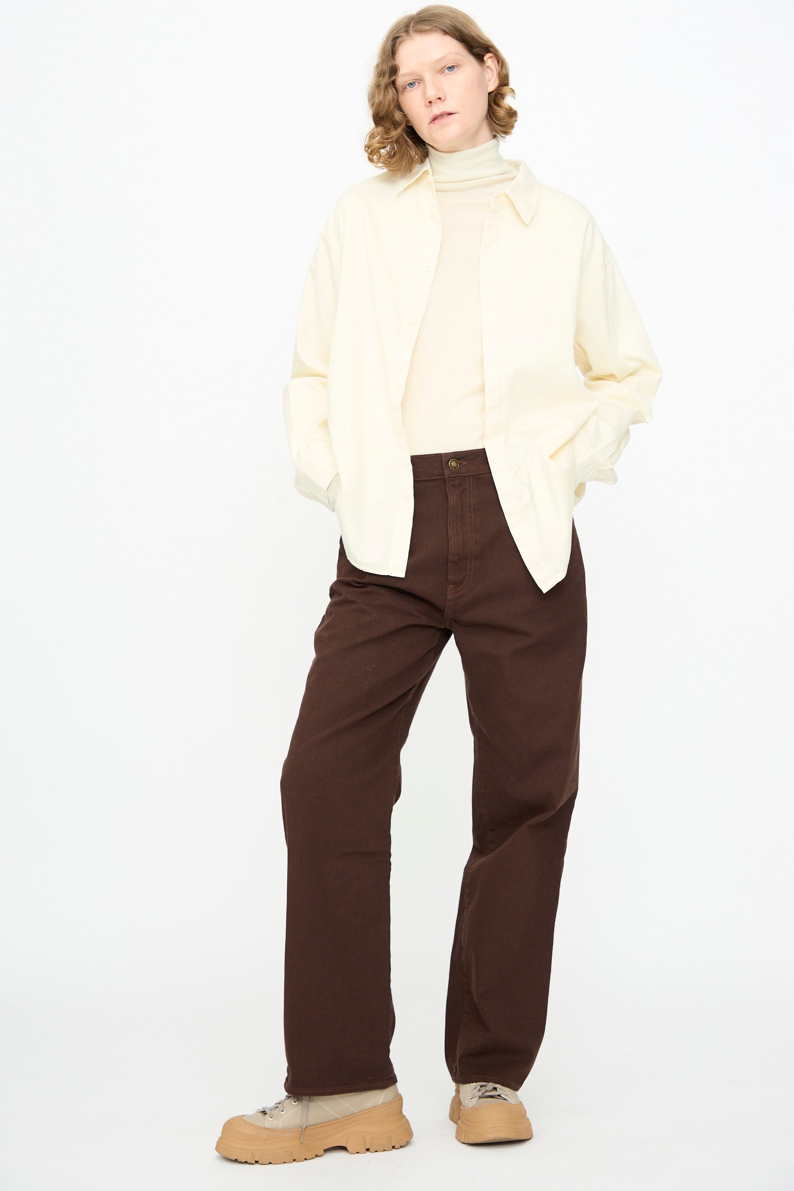 Person standing against a white background, wearing a cream turtleneck layered with the Black Crane Work Shirt in Natural, paired with brown pants and tan shoes, hands in pockets.