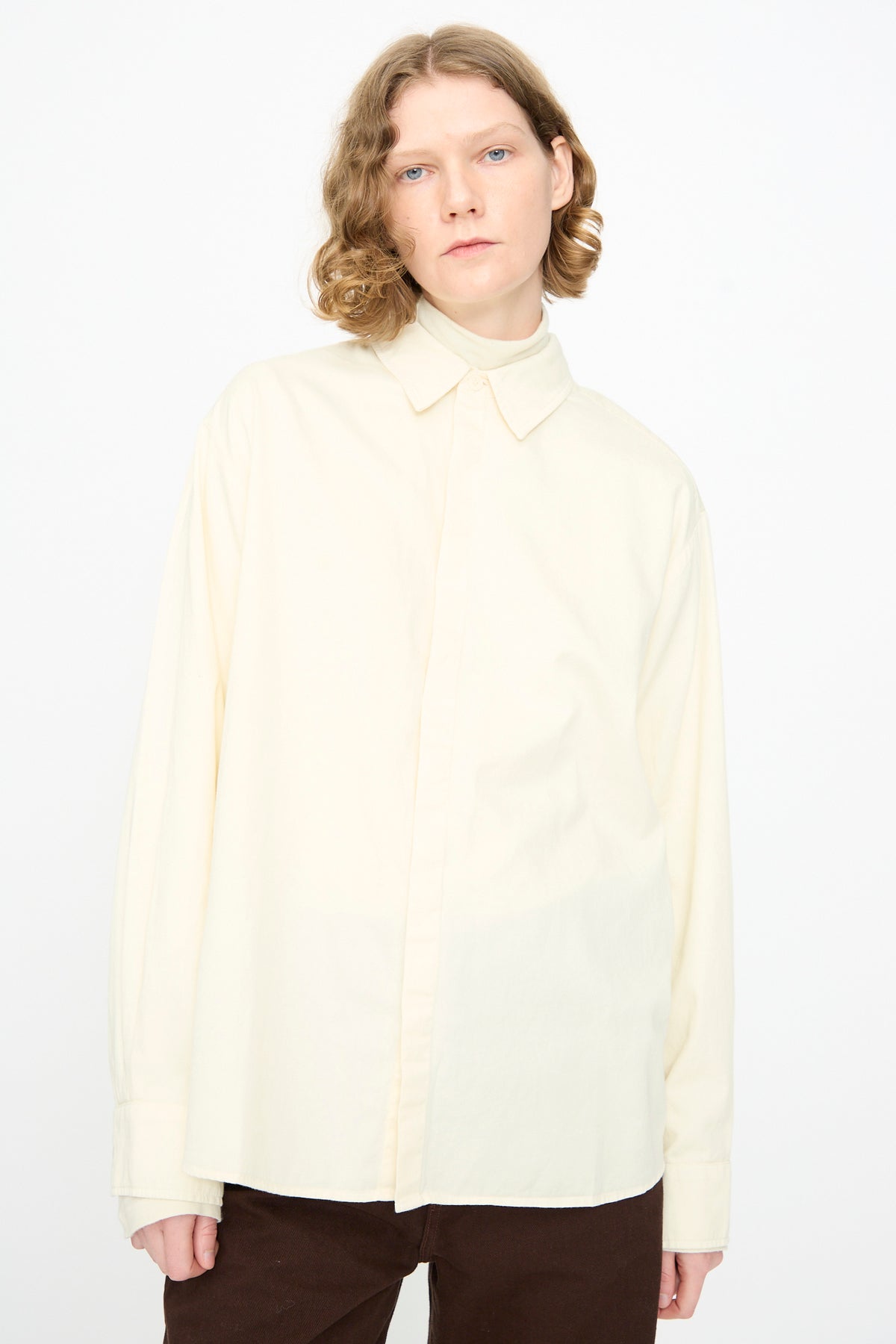 A person with shoulder-length wavy hair wears the Black Crane Work Shirt in Natural—an easy, loose-fitting cotton button-down—paired with dark pants, standing against a plain white background.