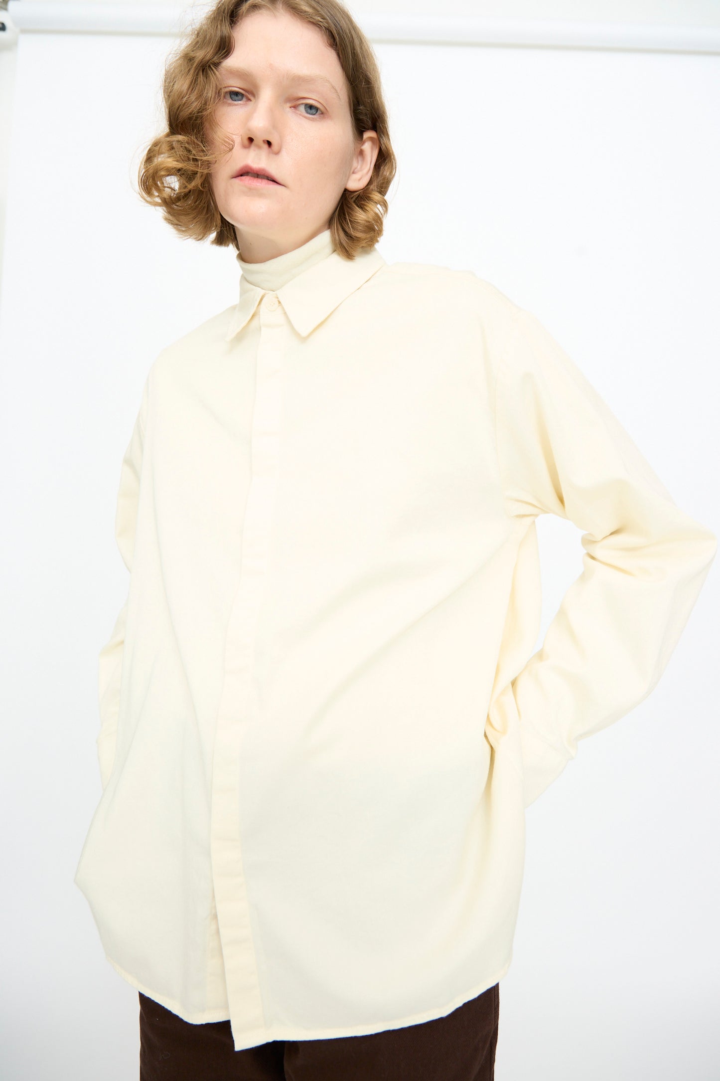 A person with short, wavy hair wears the Black Crane Work Shirt in Natural—a cream-colored, relaxed-fit cotton button down—paired with dark pants, standing against a plain white background with hands in pockets.