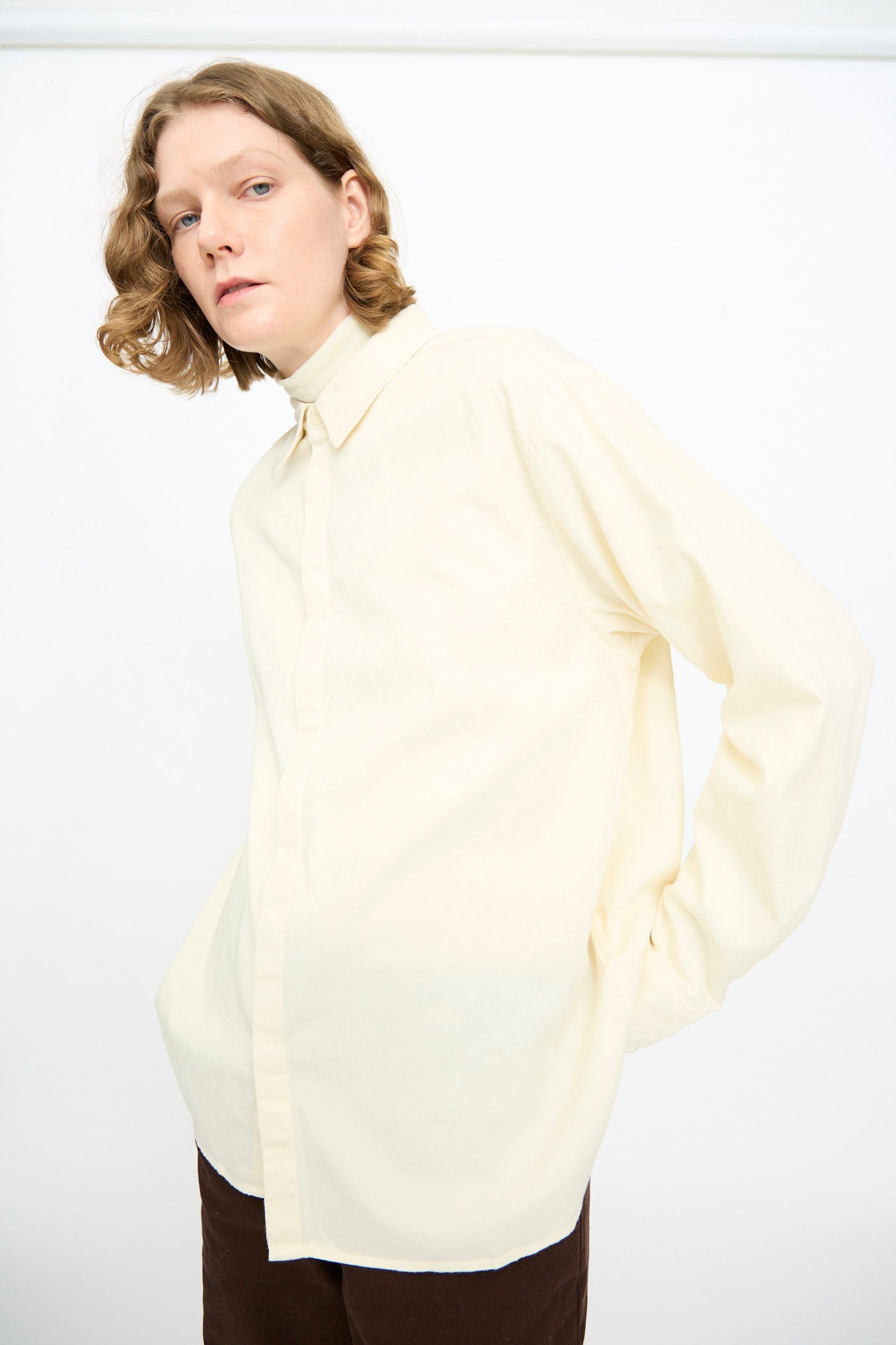A person with shoulder-length blonde hair is wearing the Black Crane Work Shirt in Natural and brown pants, standing against a plain white background.