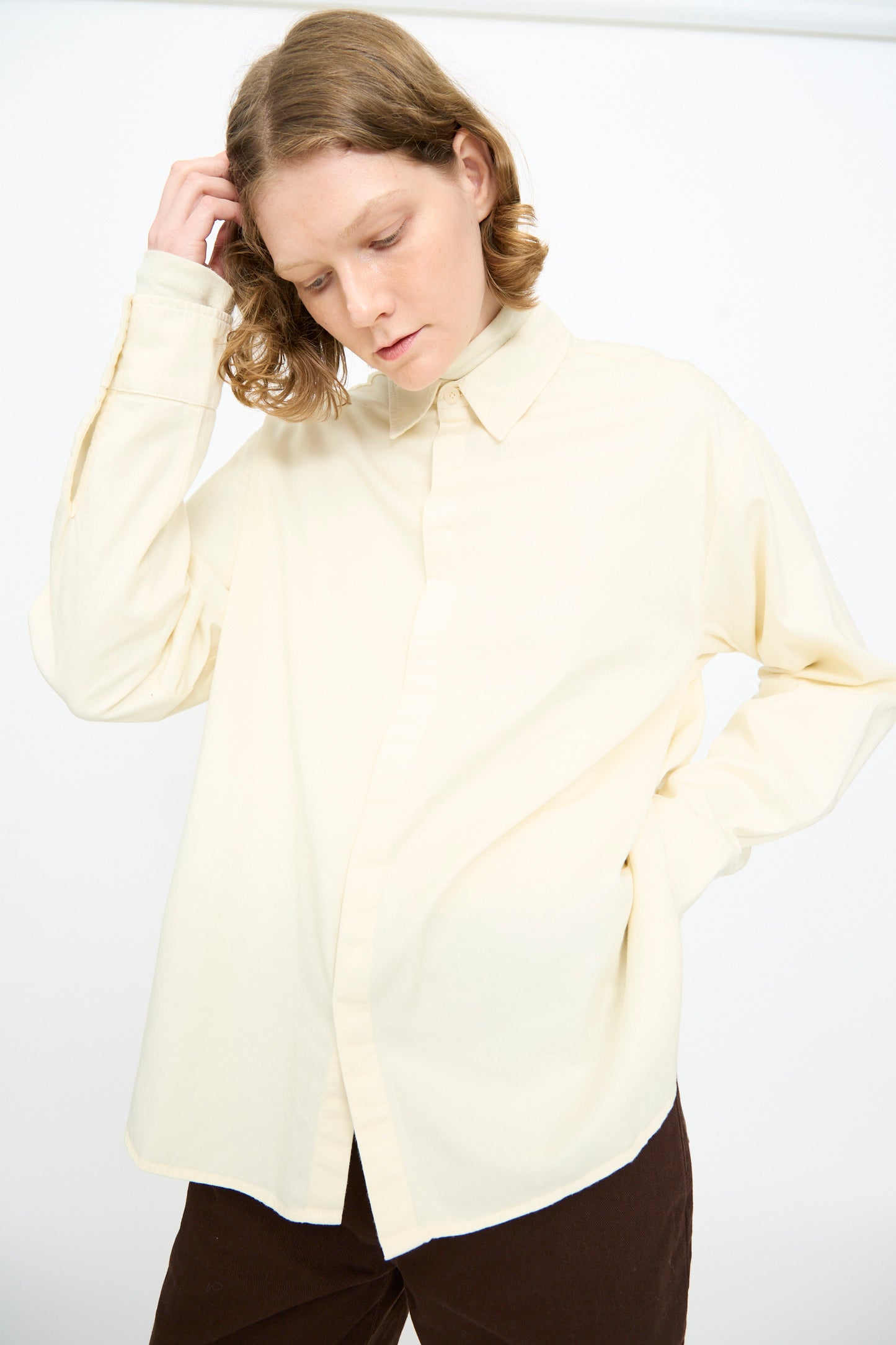 A person wearing the Black Crane Work Shirt in Natural—a loose, relaxed fit cotton button down—paired with dark pants stands against a plain white background, looking down with one hand touching their head.