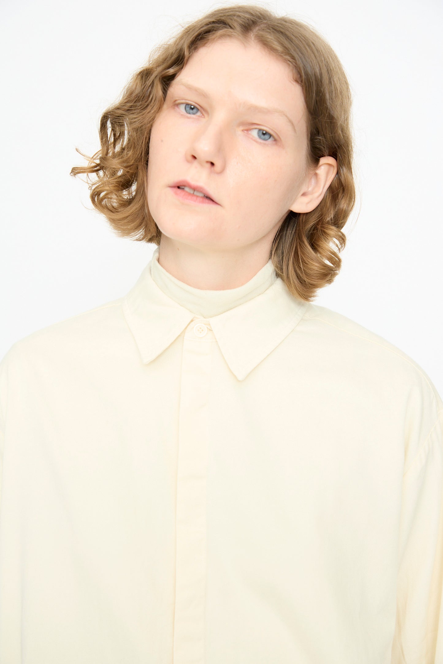 A person with wavy light brown hair stands against a plain white background wearing the Black Crane Work Shirt in Natural, buttoned up in a relaxed fit.