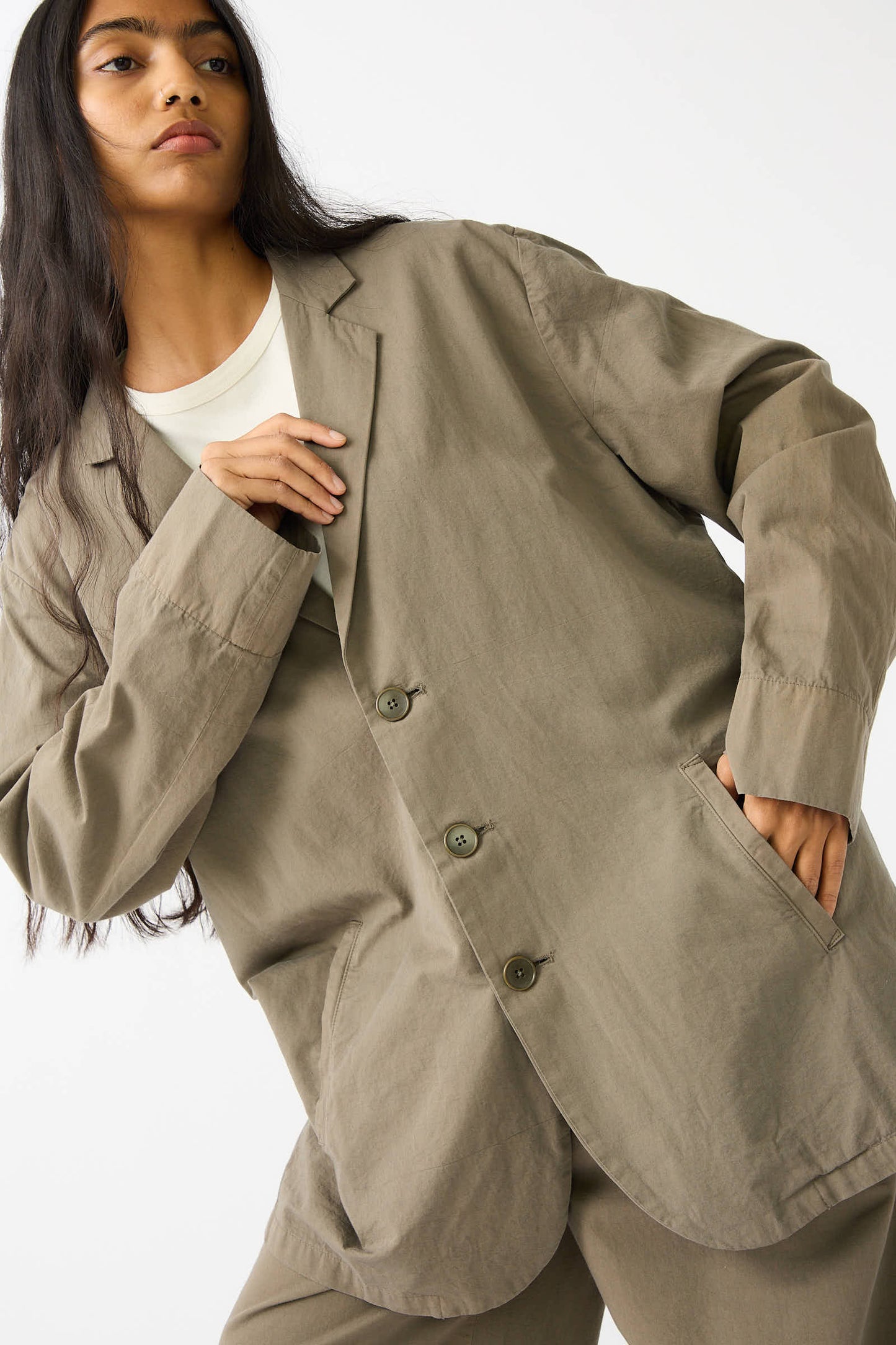 A person is wearing the Black Crane Woven Cotton Blazer in Dark Olive over a white shirt. The relaxed fit and woven Japanese cotton fabric add an elegant touch. They have long, dark hair and are posing with one hand in their pocket against a plain white background.