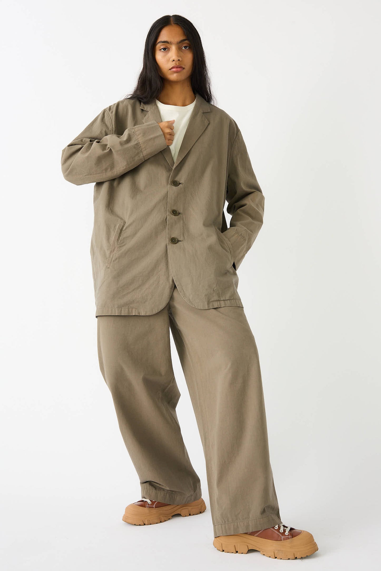 Person wearing an oversized dark olive suit with a white undershirt, posing against a plain background. They have long hair and are also wearing tan chunky shoes that complement their relaxed fit Woven Cotton Blazer in Dark Olive by Black Crane.