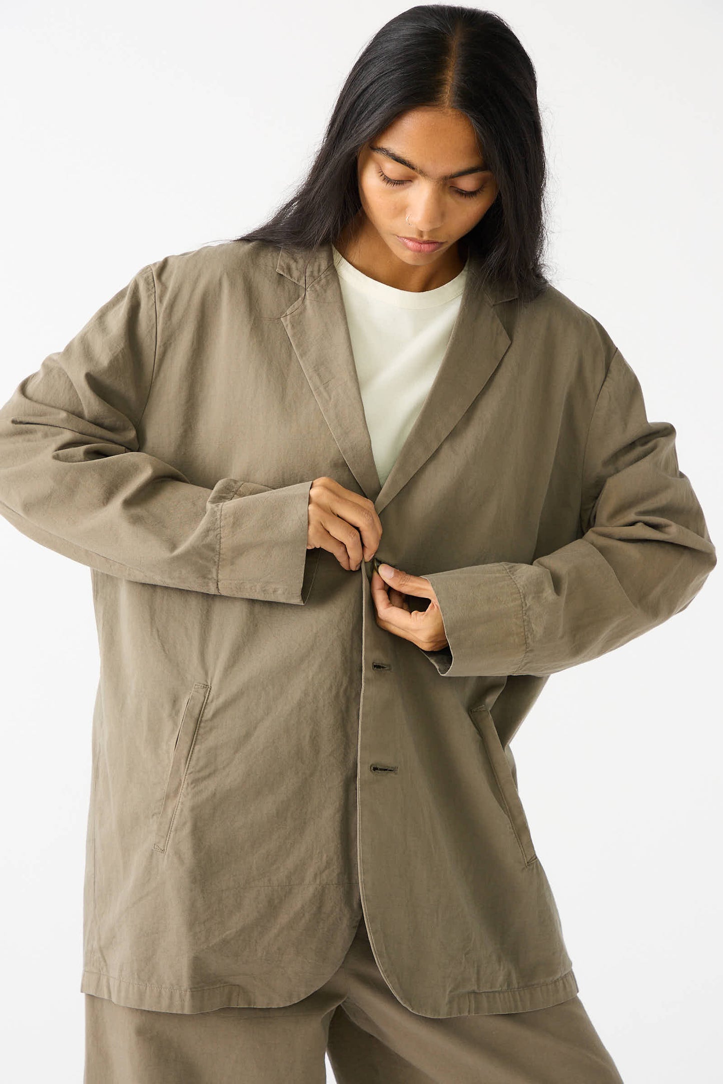 A person in a dark olive oversized blazer from Black Crane made of woven Japanese cotton, white shirt, and beige pants, buttoning the jacket. The individual has long, straight, dark hair and is standing against a plain white background.