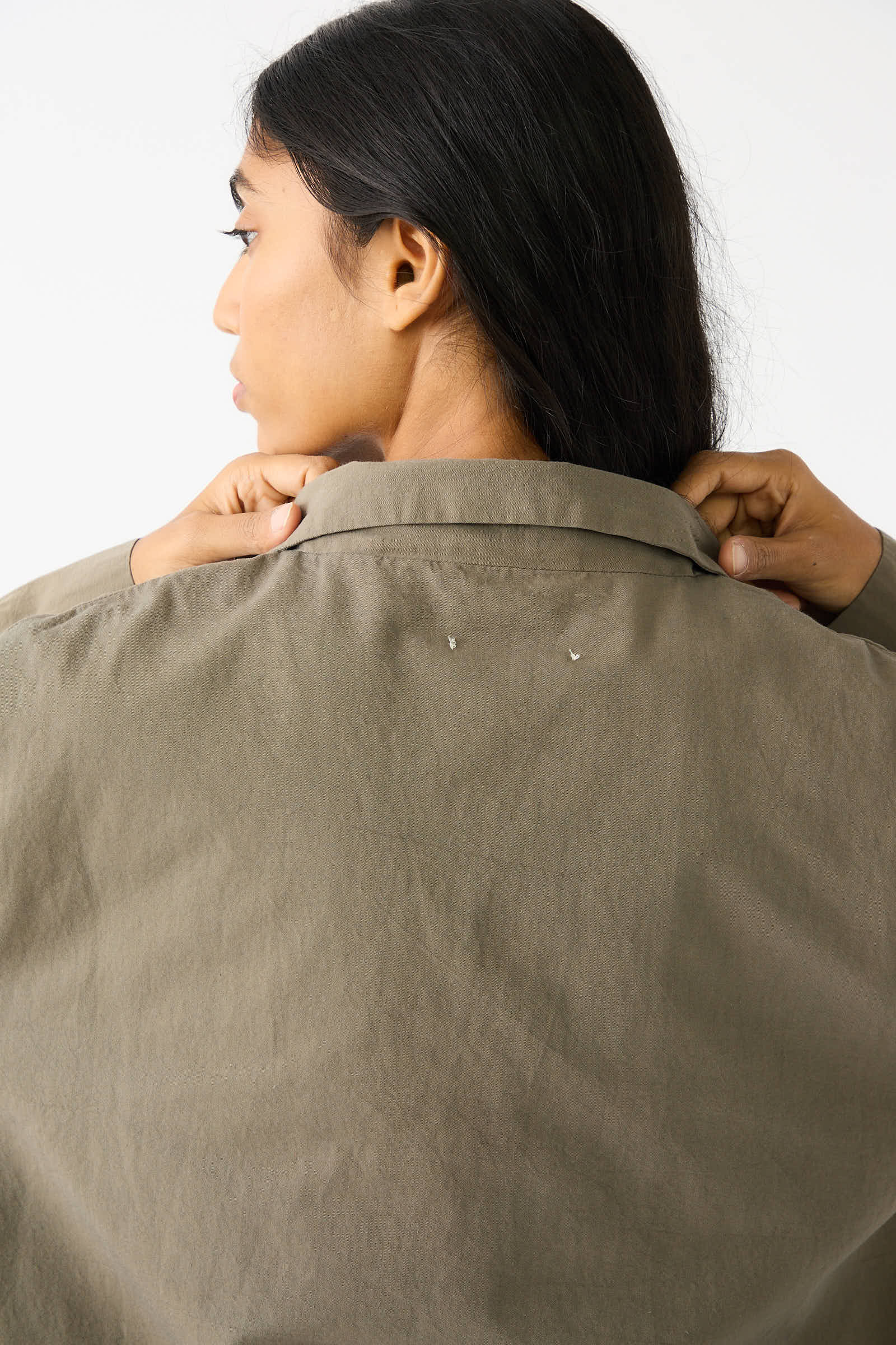 A person with long dark hair, facing away, adjusts the collar of a Woven Cotton Blazer in Dark Olive by Black Crane.