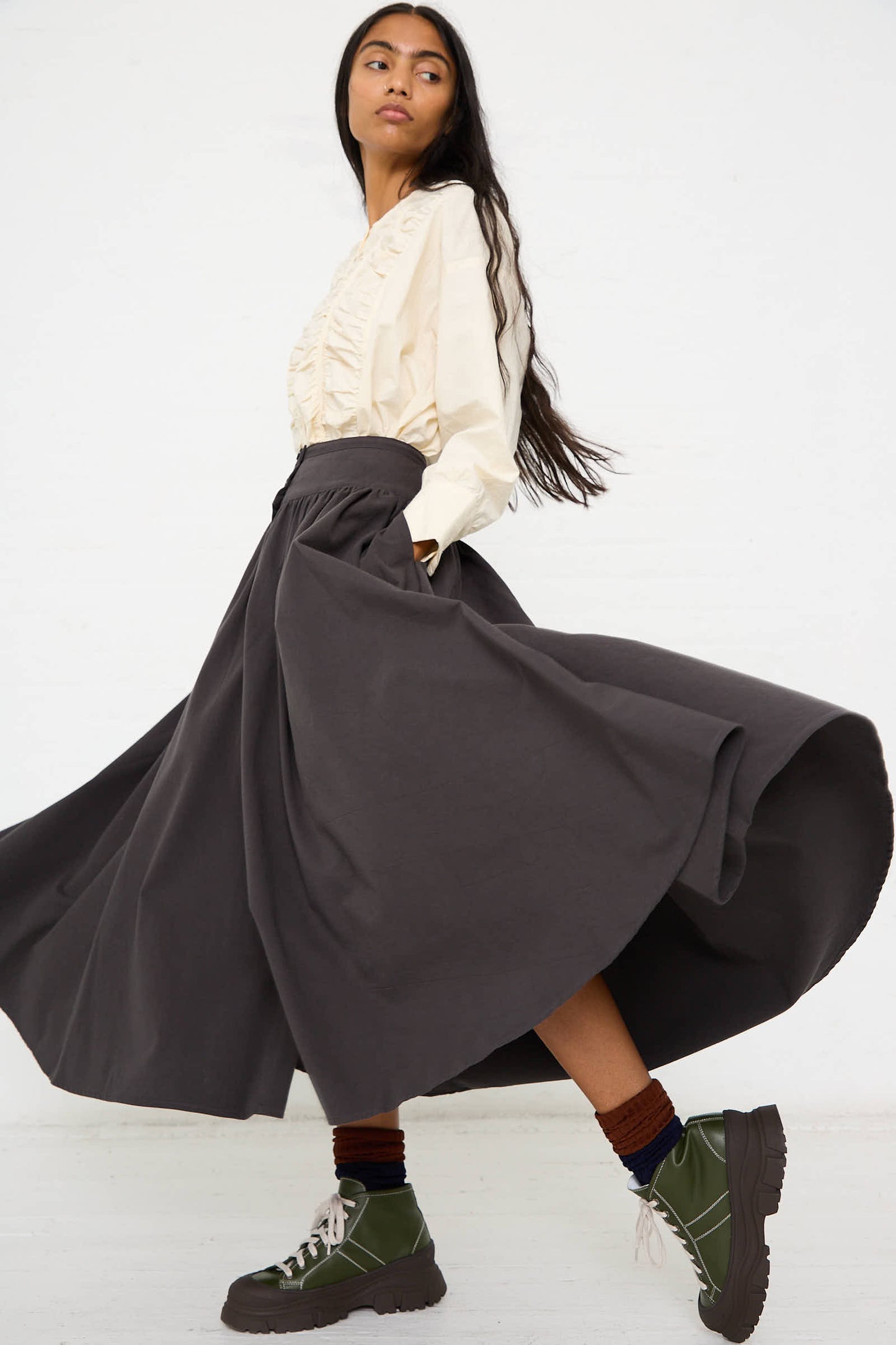 A woman with long hair wears a cream blouse and a Woven Cotton Circular Skirt in Dark Grey by Black Crane, paired with chunky green shoes. She is captured mid-stride in side profile.